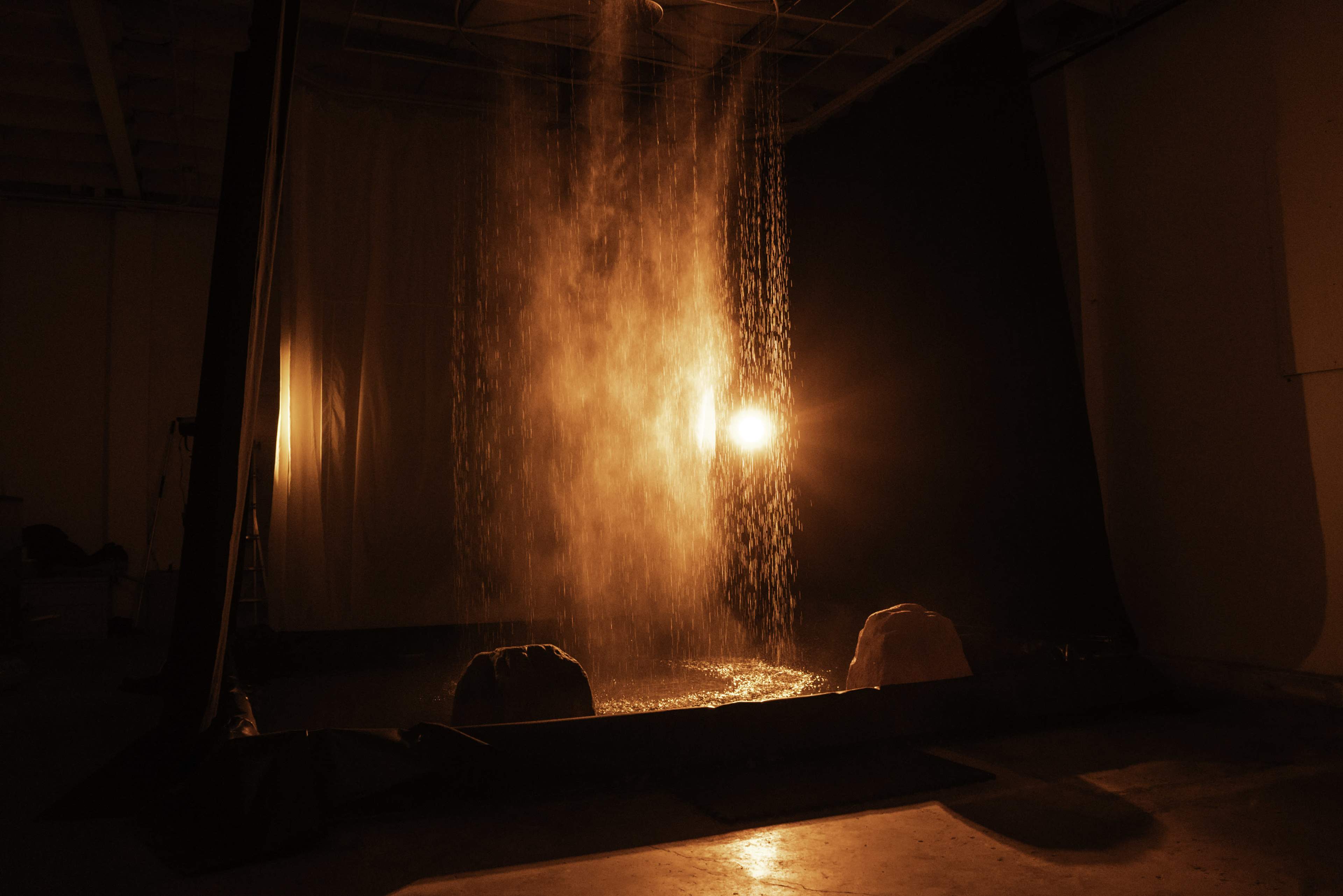 A cascade of water falls in a dimly lit studio, illuminated by a bright orange light source.