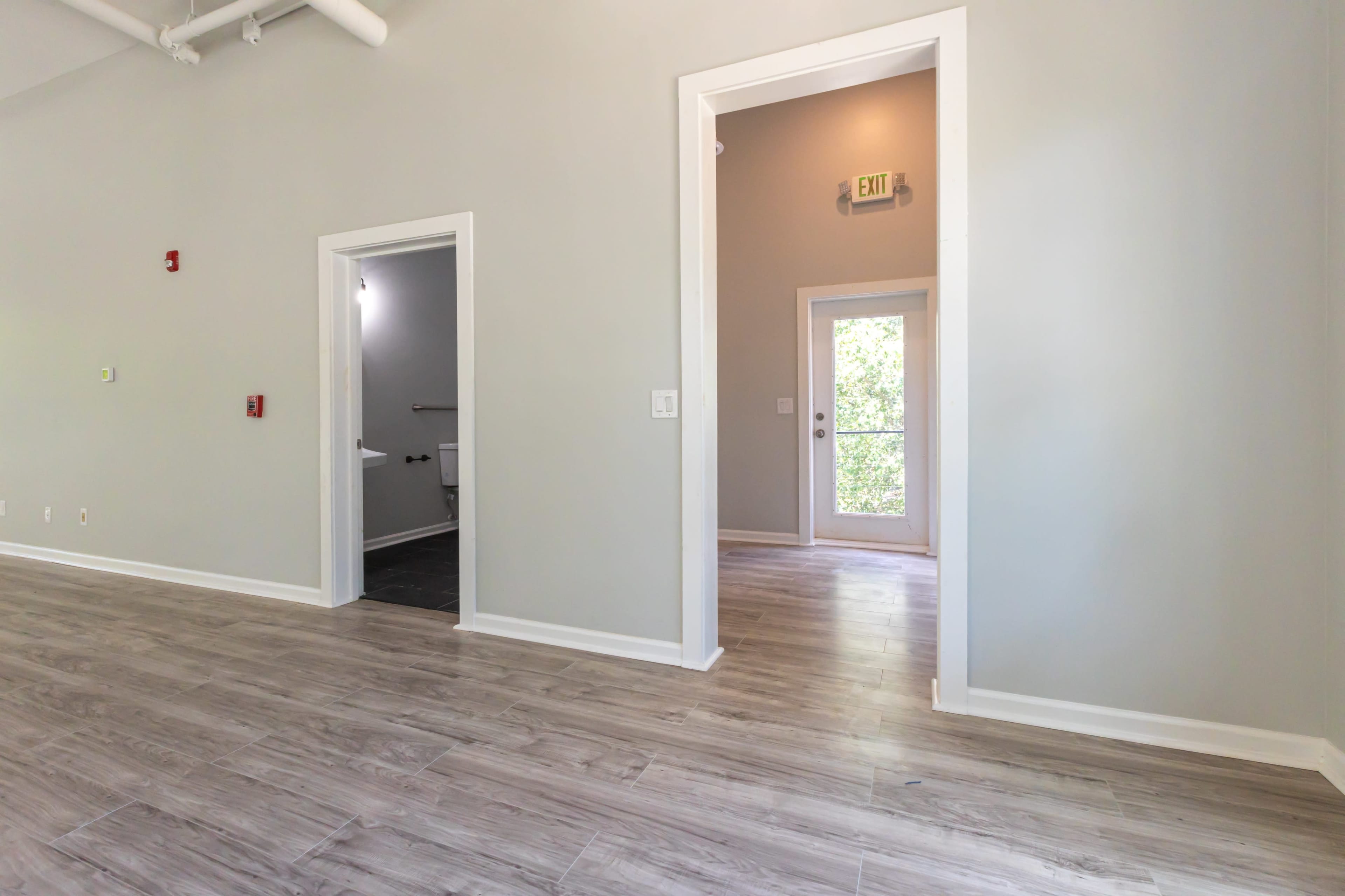 The image shows a spacious, light-colored room with wooden flooring, featuring doorways leading to other rooms and a view of a door opening to the outdoors.