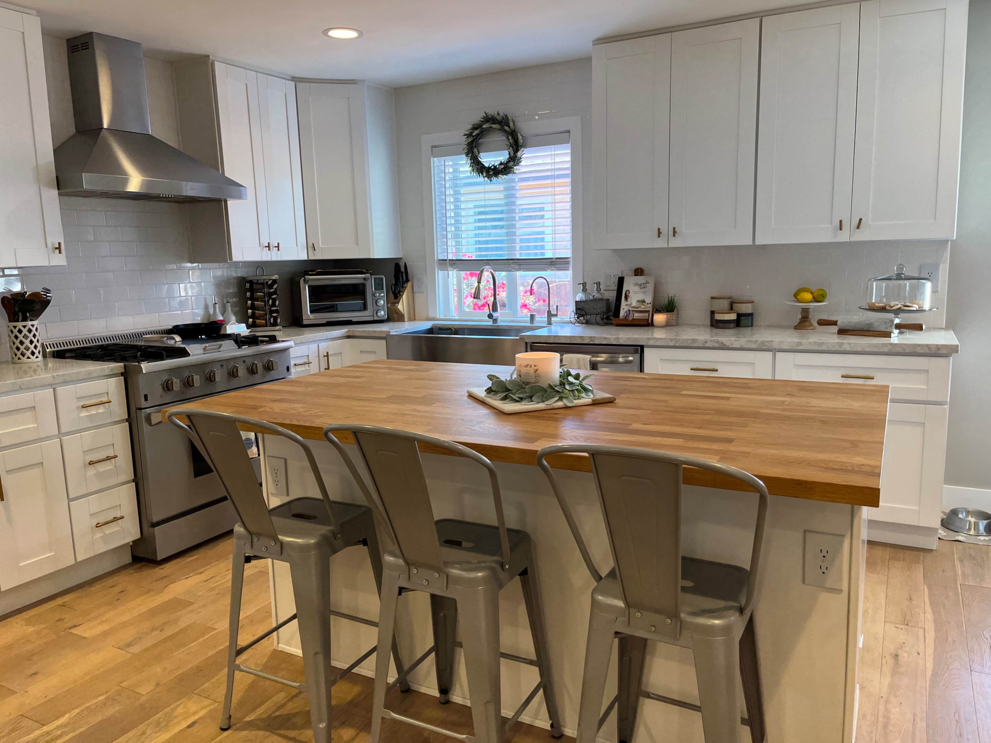 A modern kitchen features white cabinets, a central wooden island with metal stools, and a window overlooking a garden.