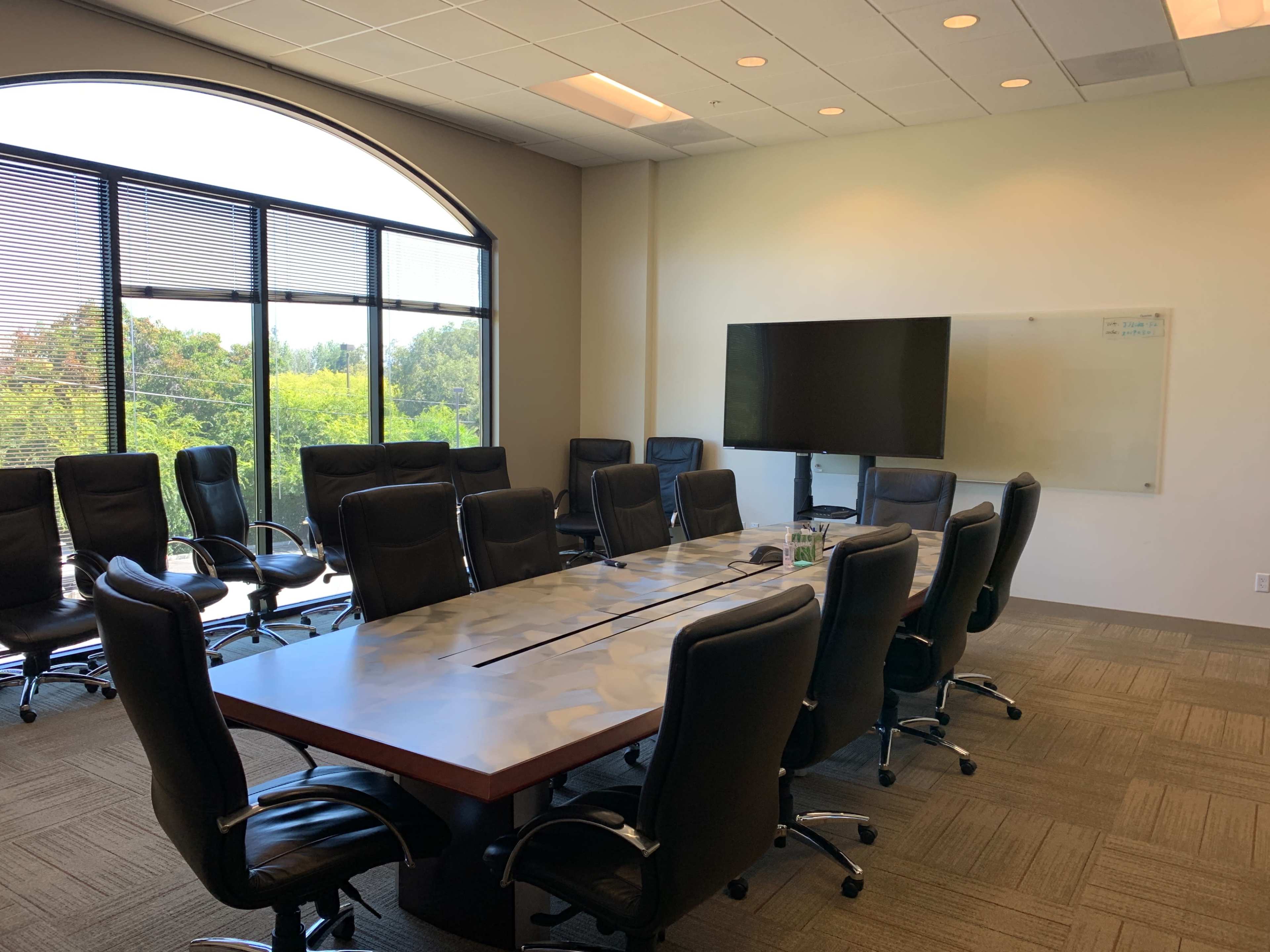 A large conference room with a long table surrounded by black chairs, a television mounted on the wall, and a whiteboard.