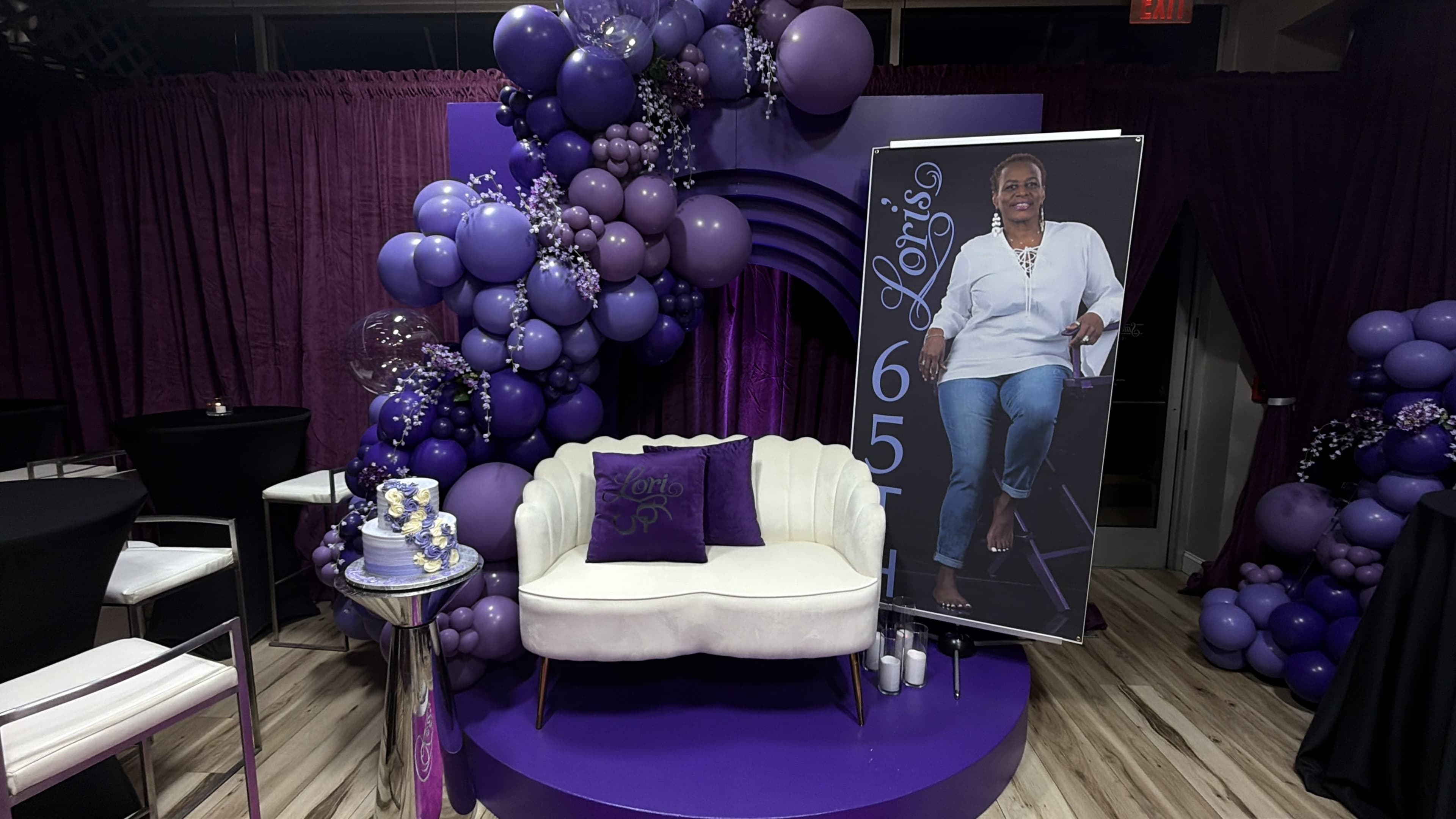 The image features a purple-themed seating area with a white couch, a large balloon arrangement, and a tall poster of a woman, all set against a purple backdrop.