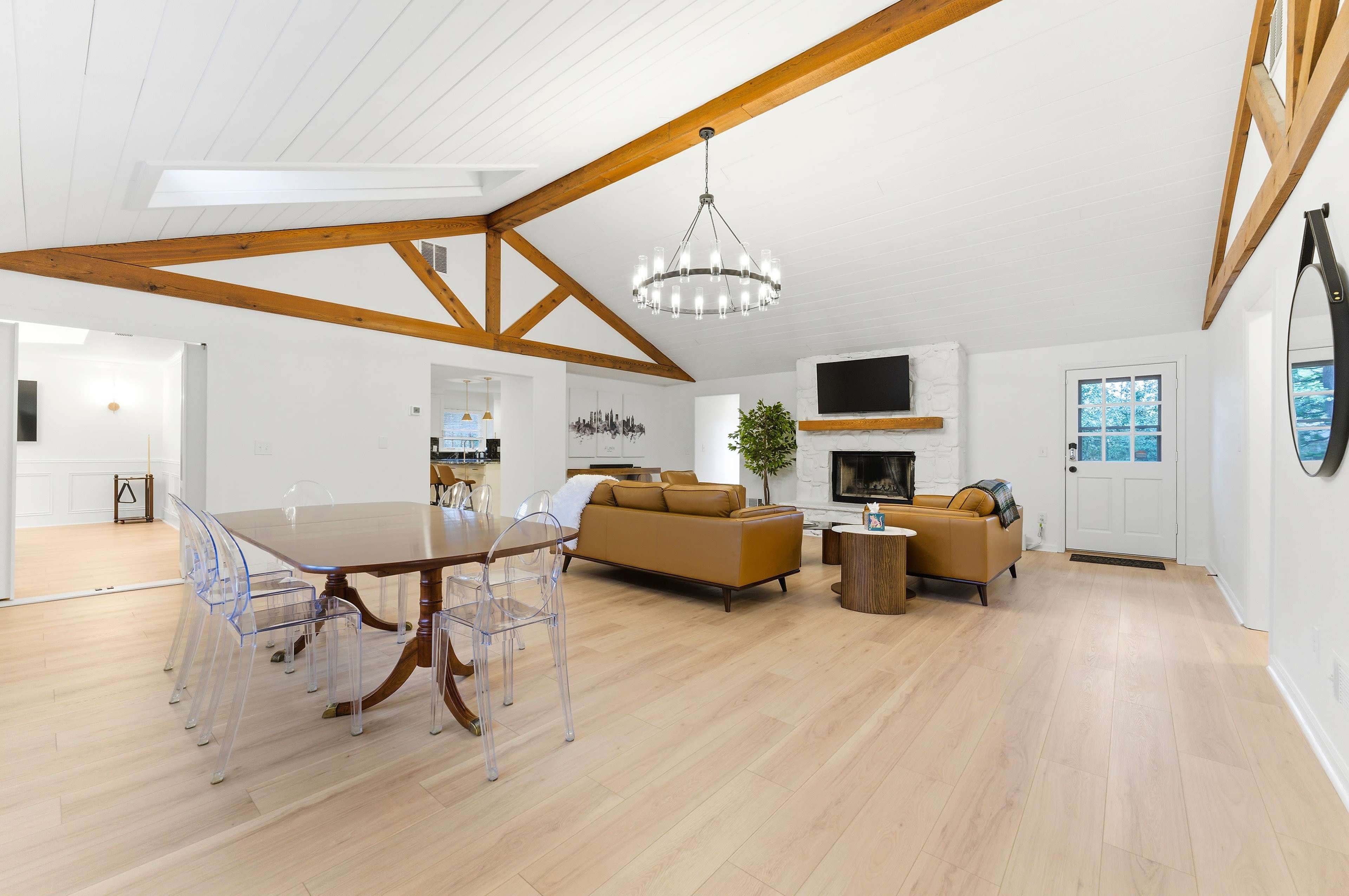 The image depicts a spacious living area featuring wooden beams, a large dining table, comfortable seating, and a fireplace.