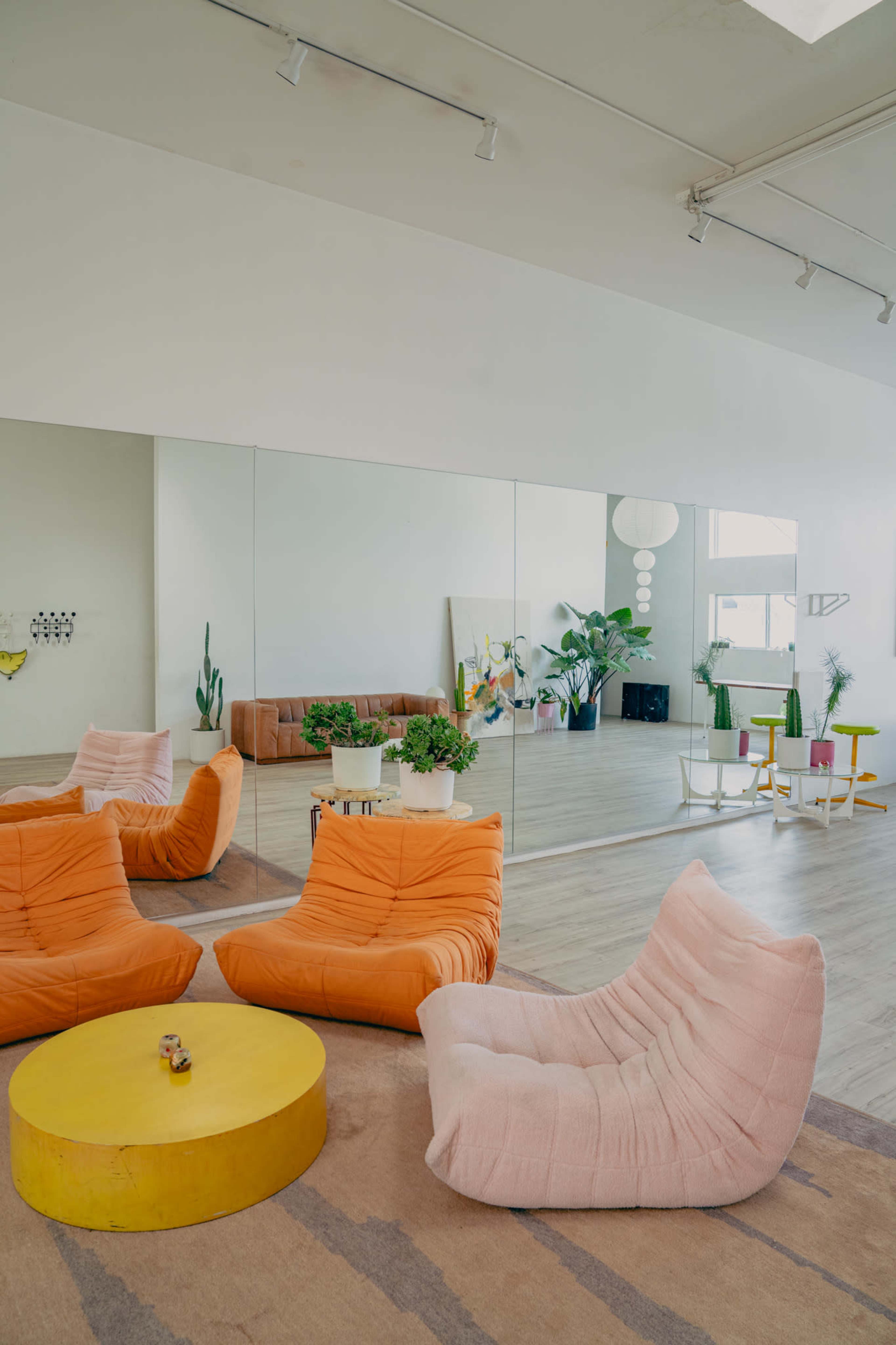 The image shows a modern living space featuring two bright lounge chairs, a round yellow coffee table, and large mirrors reflecting additional seating and decorative plants.