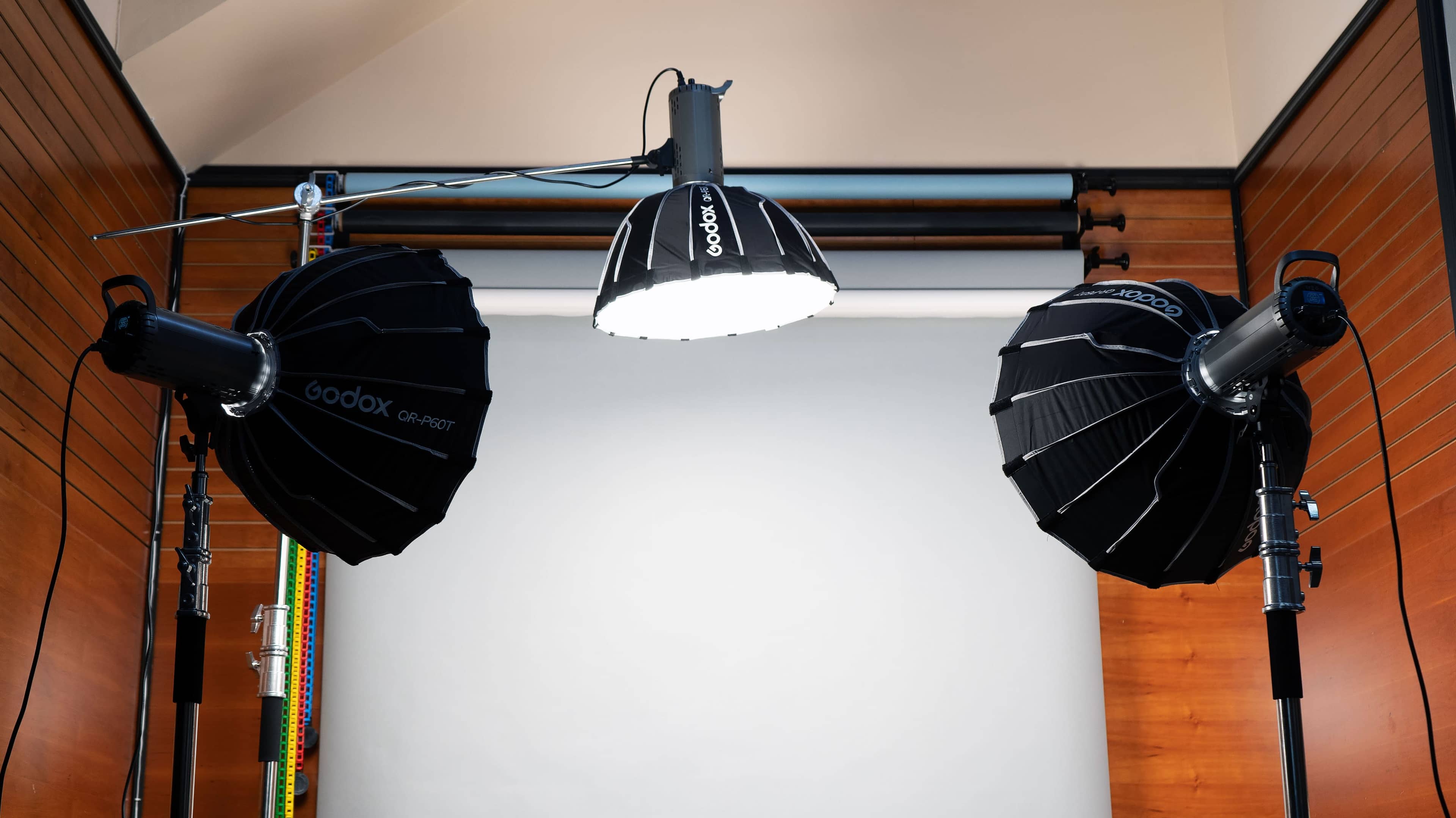 A photography studio setup with three Godox lights mounted on stands, focused on a plain backdrop.