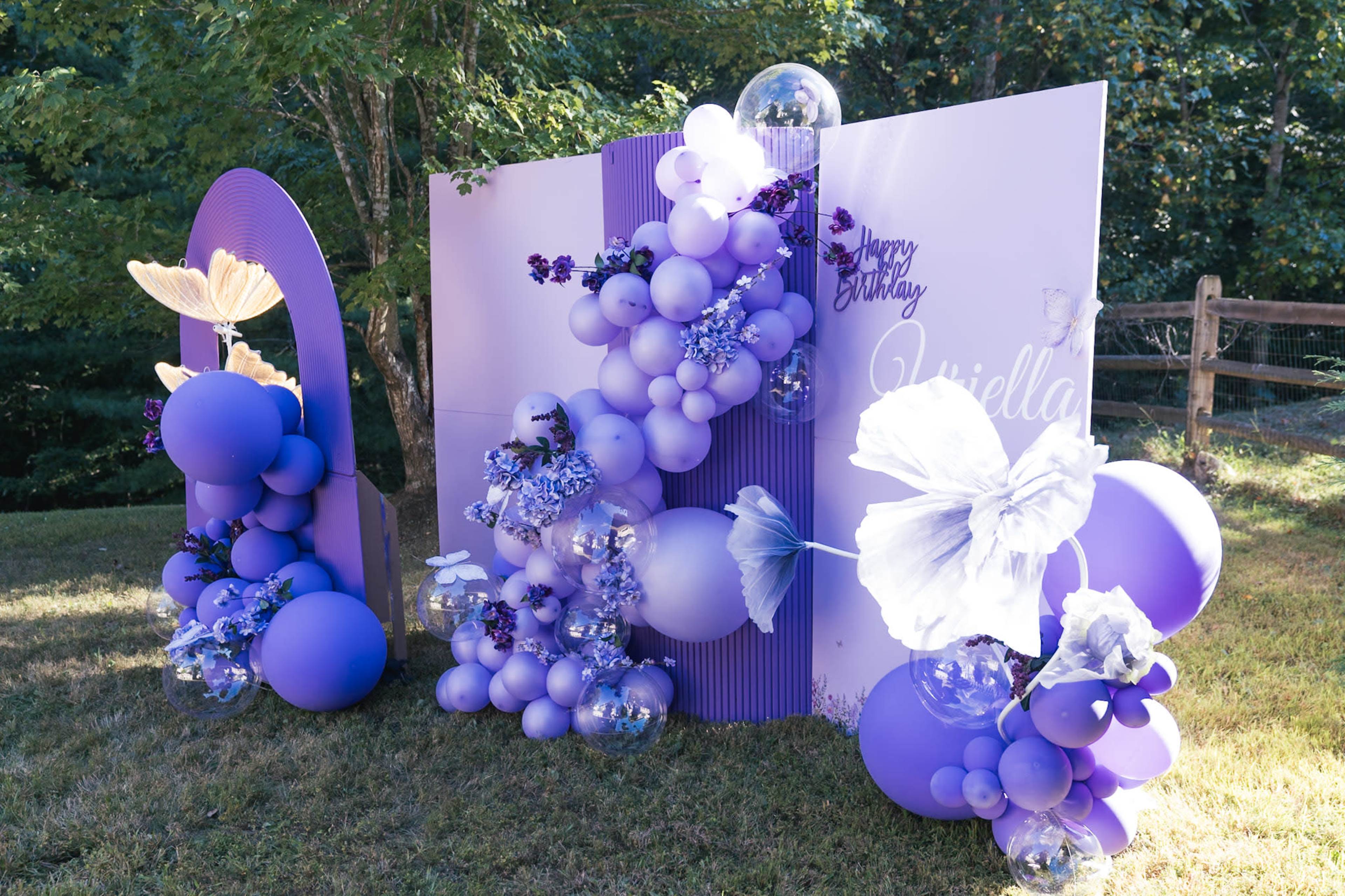 A colorful birthday backdrop featuring purple balloons, flowers, and a large birthday greeting is set in a grassy outdoor area.