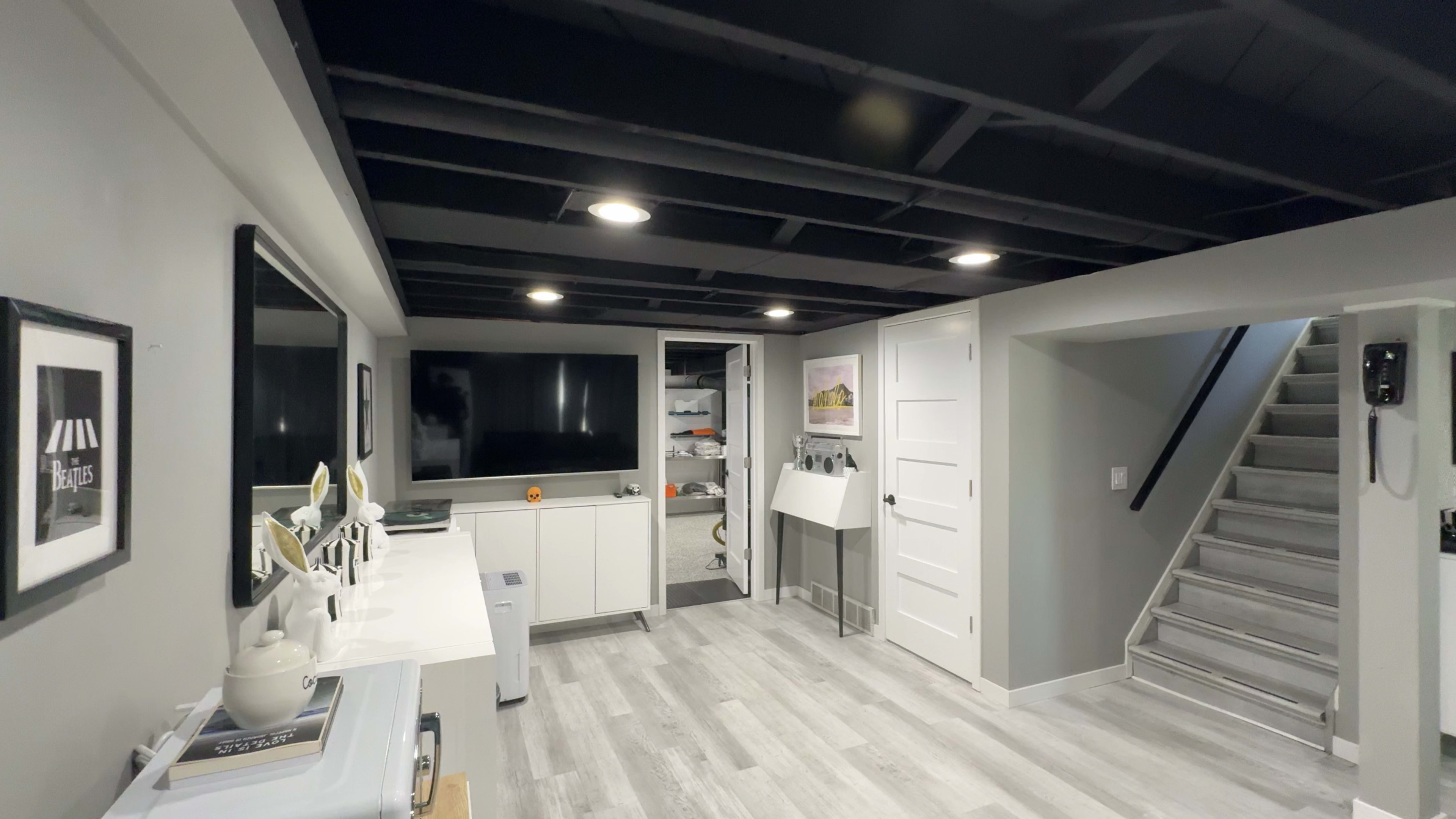 The scene shows a modern basement area featuring a TV, storage cabinets, a small sink, and a staircase leading to another level.