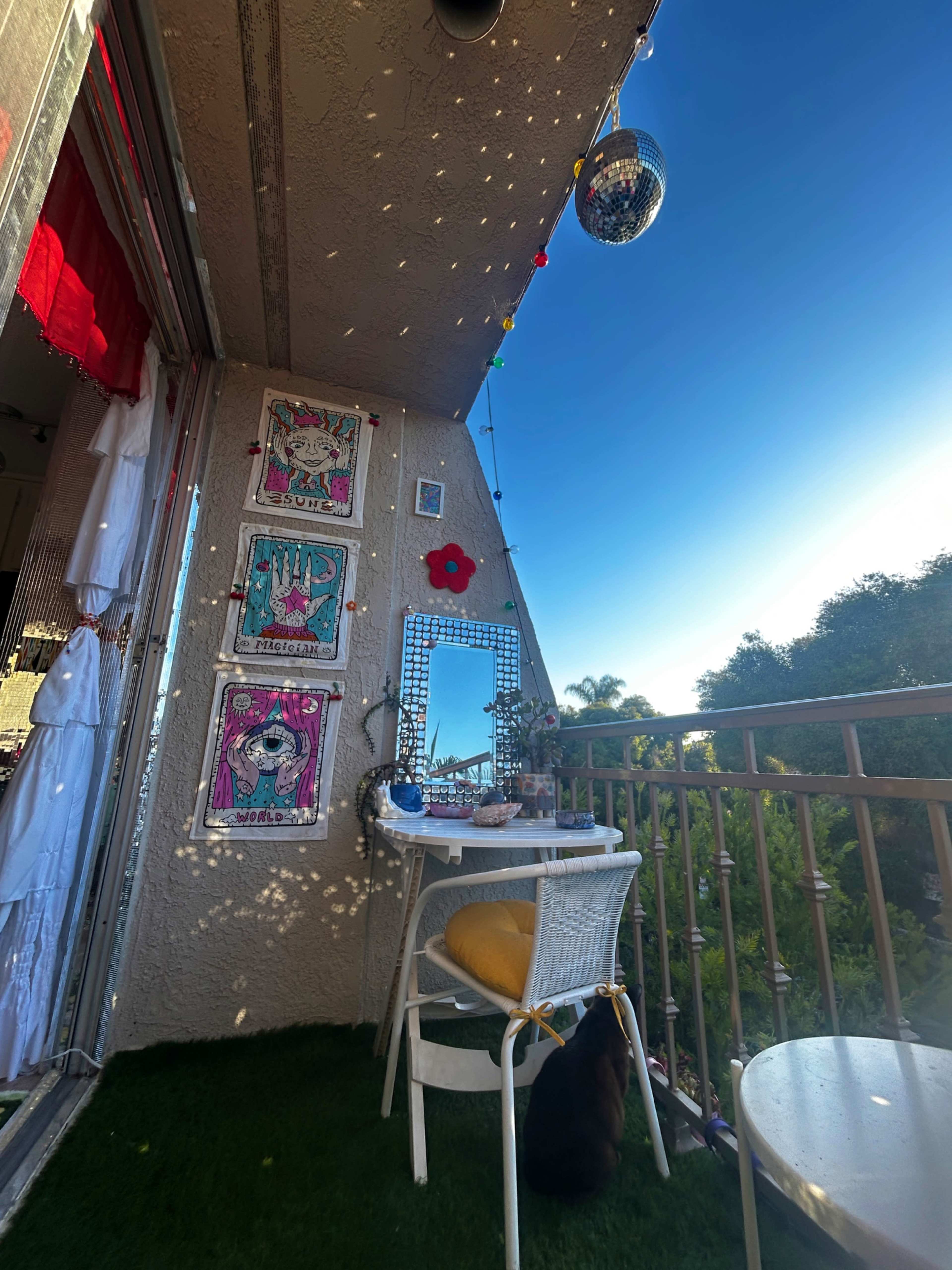 A small balcony features a table and chair with decorative artwork on the wall, surrounded by plants and a bright blue sky.