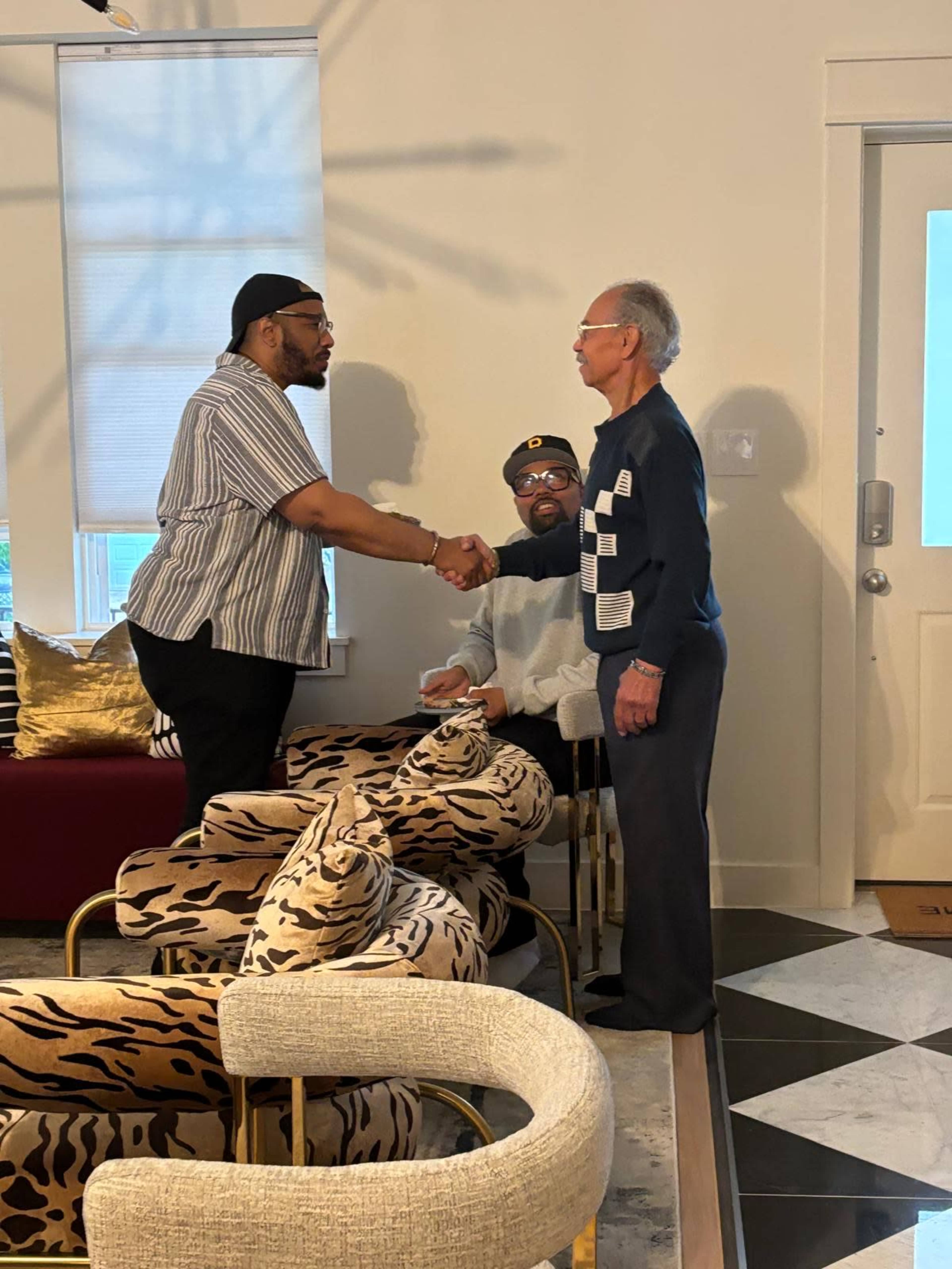 Three men are interacting in a living room, with one man shaking hands with another while a third man sits nearby.
