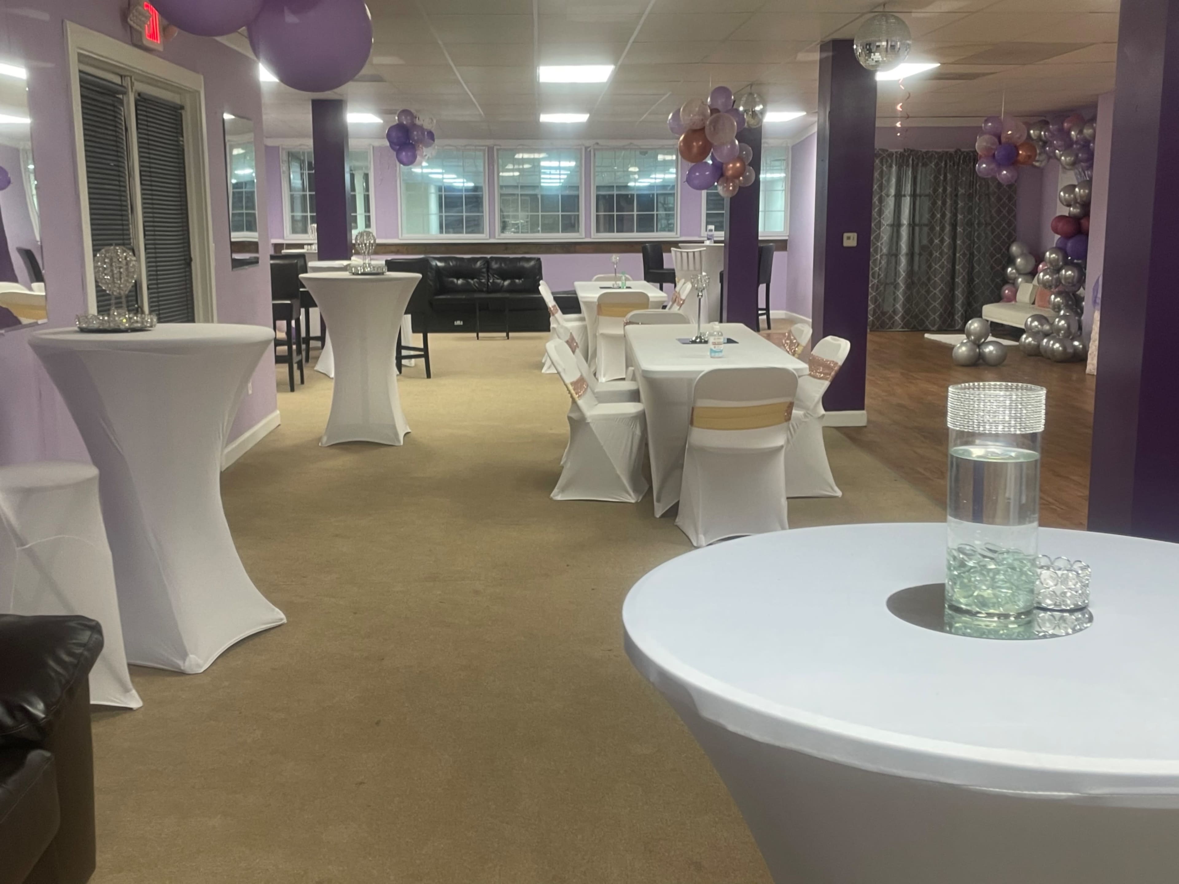 A party venue with tables dressed in white covers, purple and silver decorations, and a spacious area filled with balloons and a mirrored backdrop.