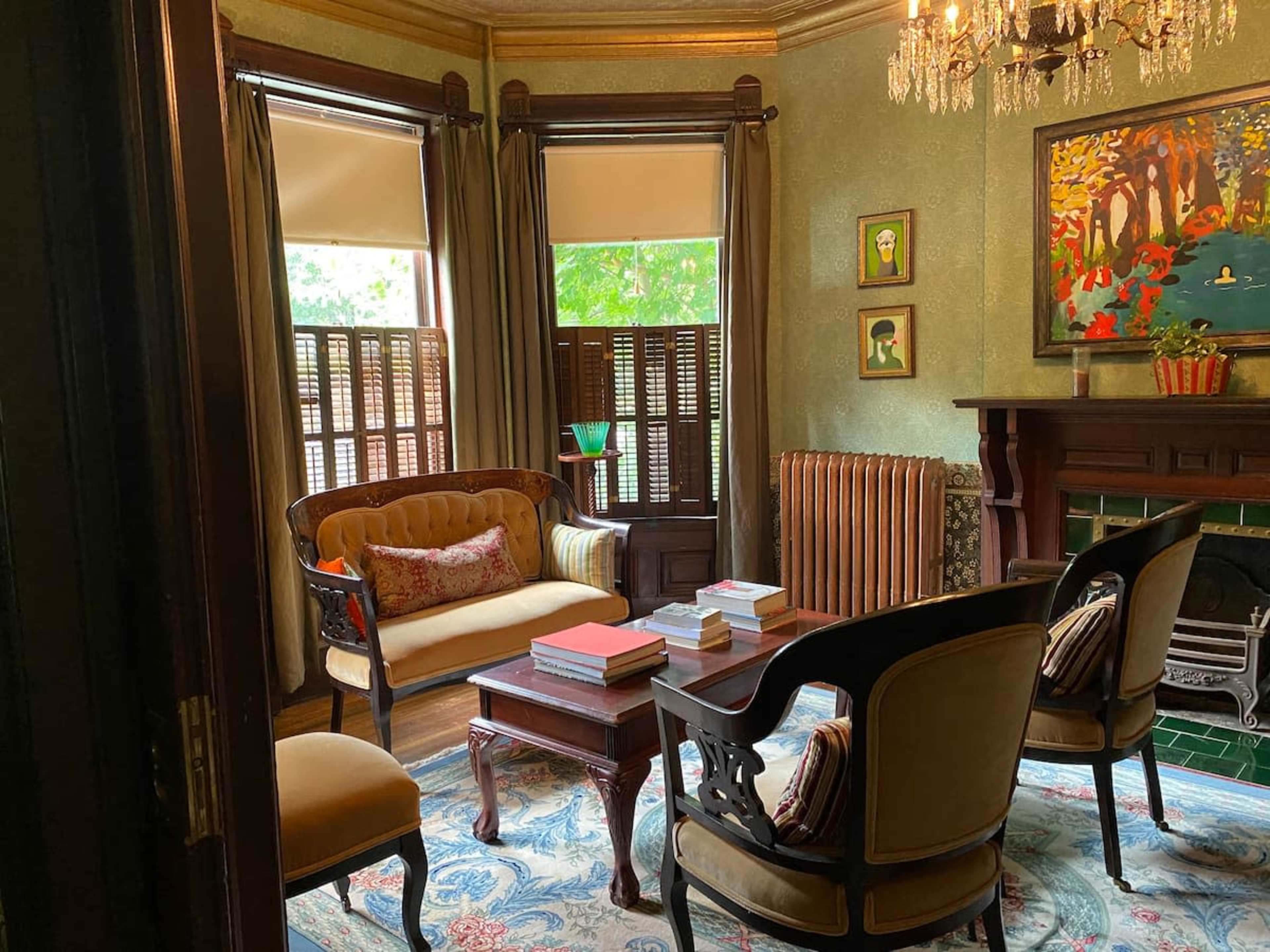 A cozy sitting room features a vintage sofa, armchairs, a wooden coffee table, and large windows with blinds.