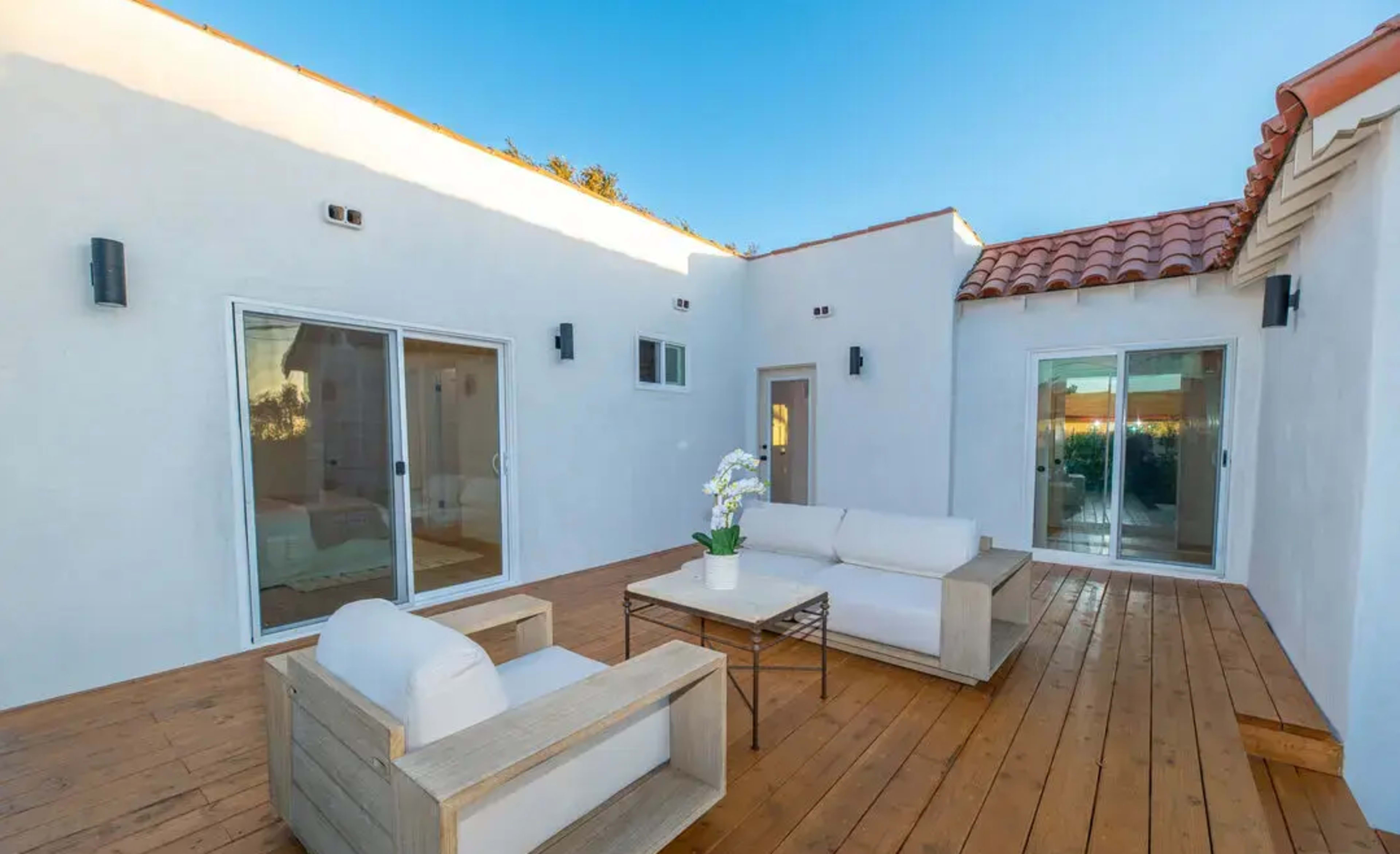 The image shows a sunlit outdoor patio area with white furniture, wooden flooring, and sliding glass doors leading to an interior space.