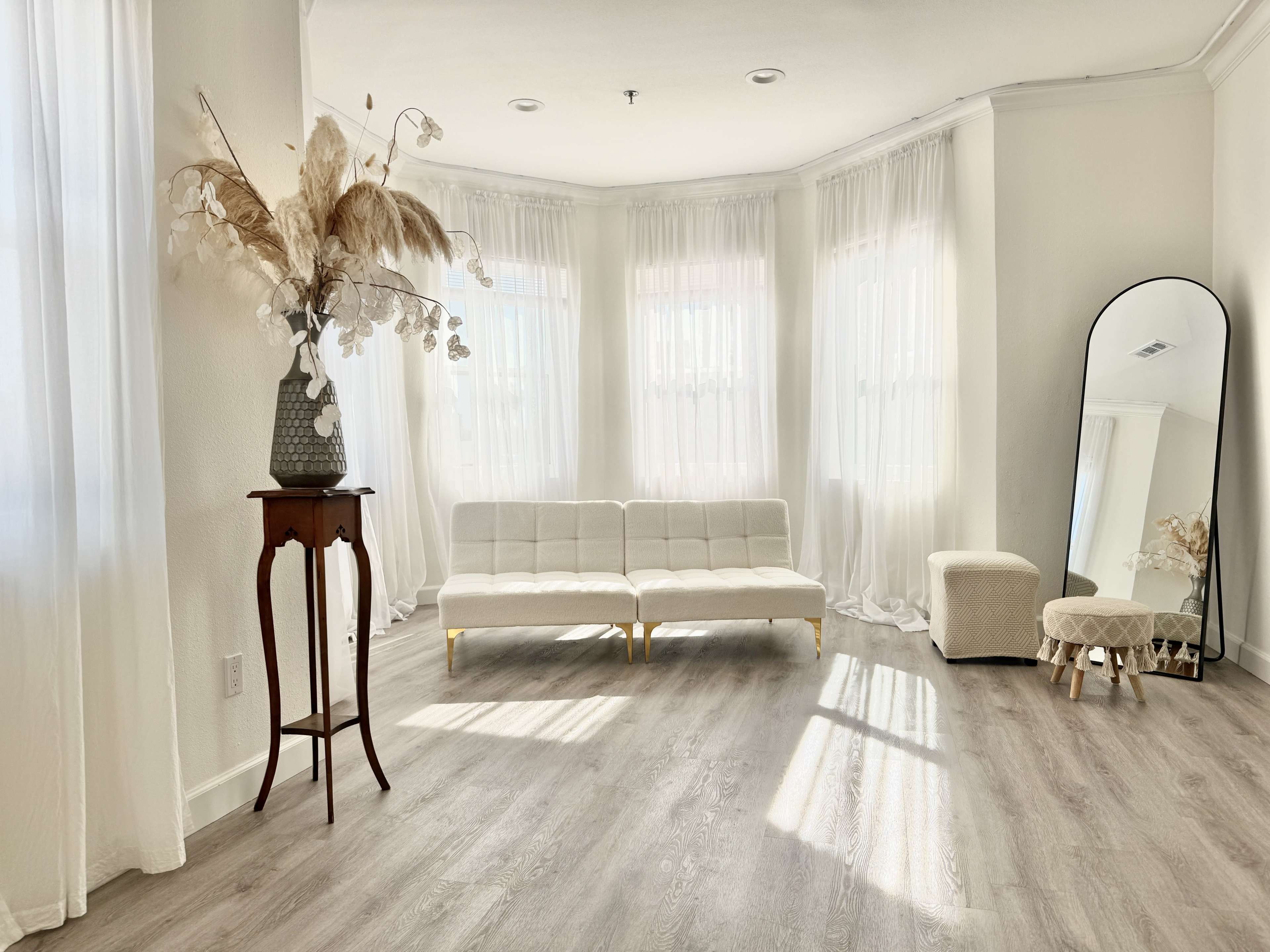 The image shows a bright and airy living room with large windows, a white couch, a mirror, and decorative elements on a small table.