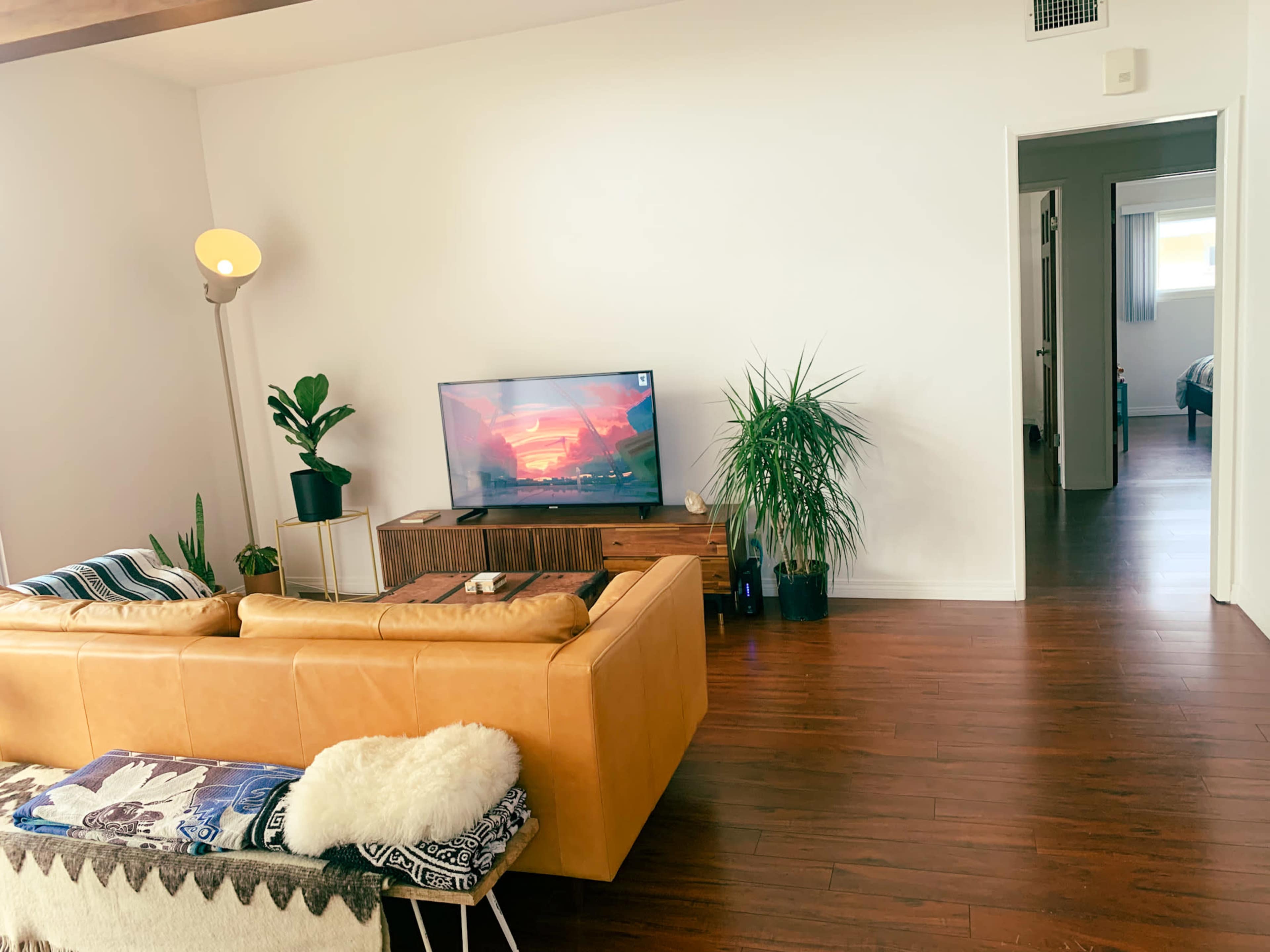 A cozy living room features a beige leather couch, a wooden TV stand with a television displaying a colorful scene, and potted plants alongside a doorway leading to another room.
