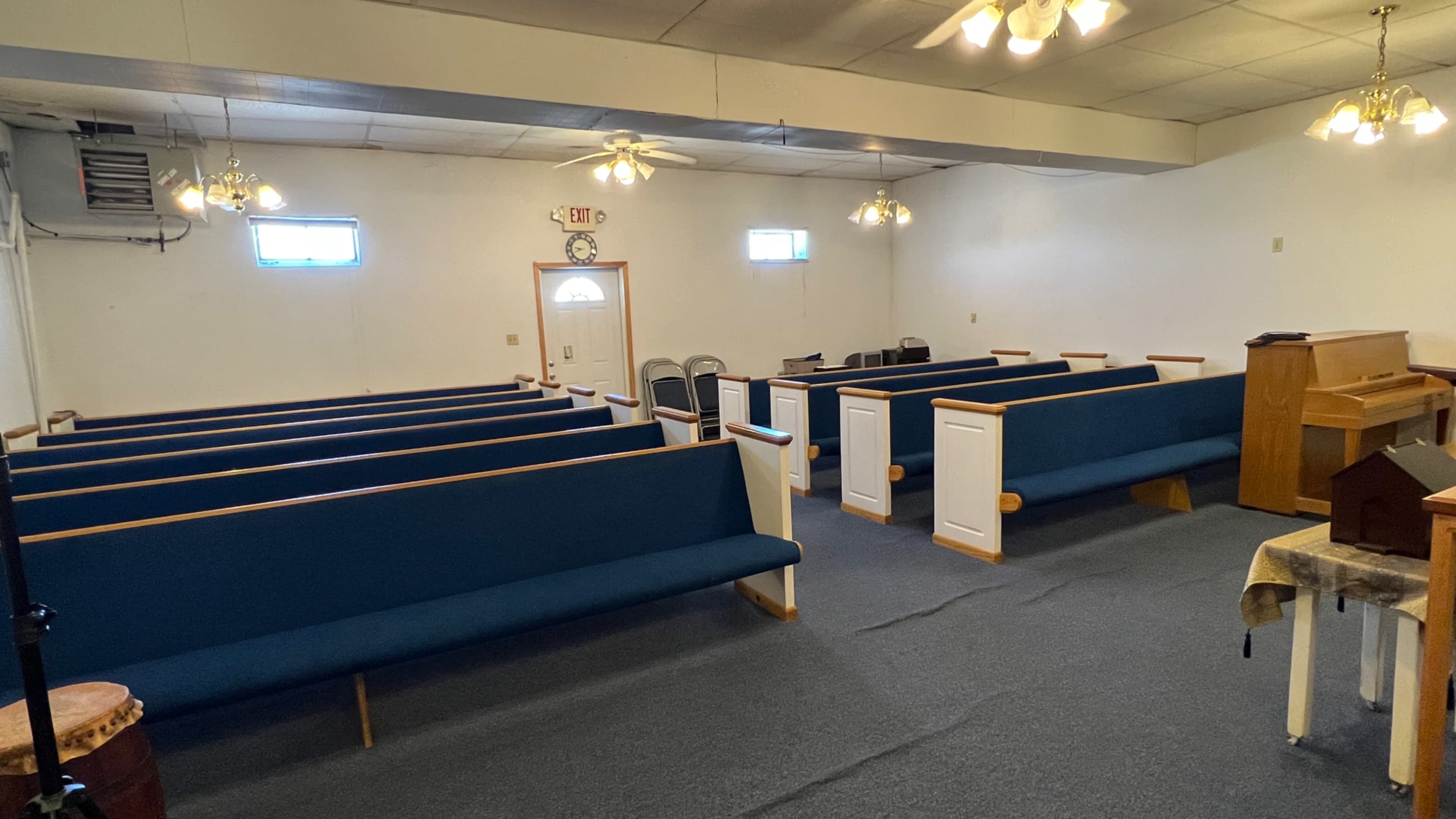An interior view of a small church or chapel with rows of blue pews, a piano, and a simple altar area.