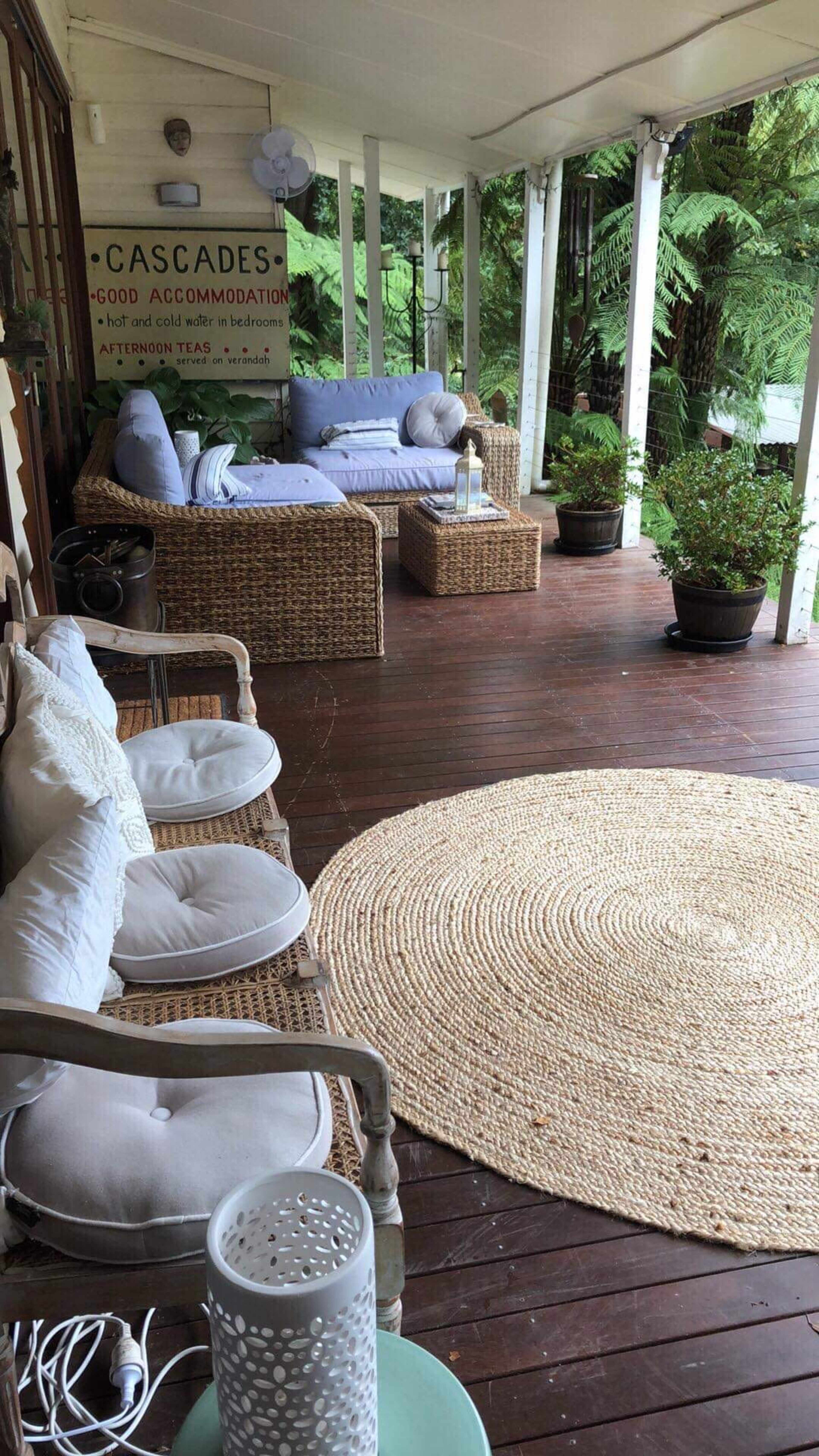 A covered porch area furnished with wicker seating, a round rug, and potted plants, set amidst greenery.