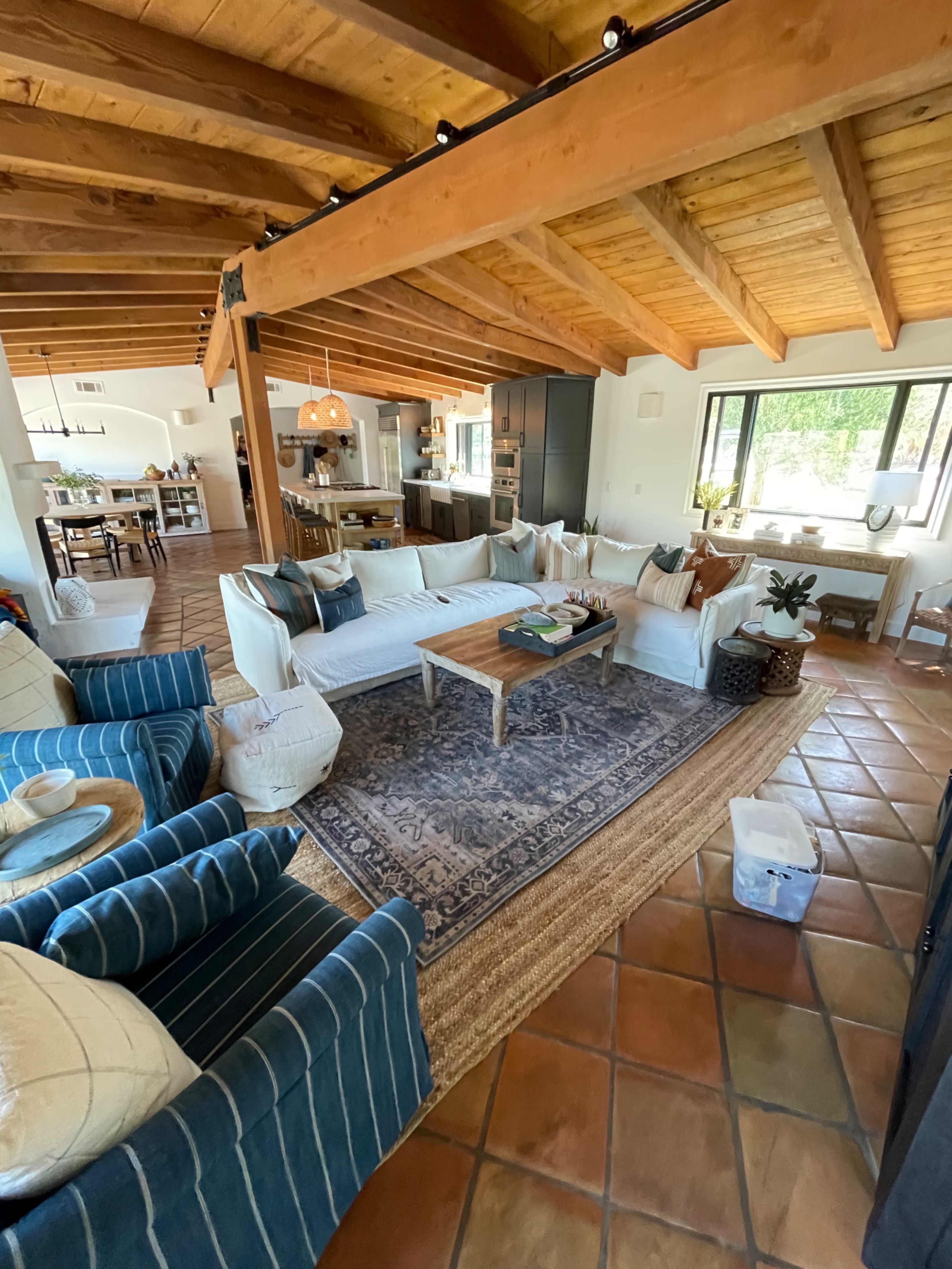 A spacious living room features a large sectional sofa, two striped chairs, a wooden coffee table, and tiled flooring, with exposed wooden beams overhead.