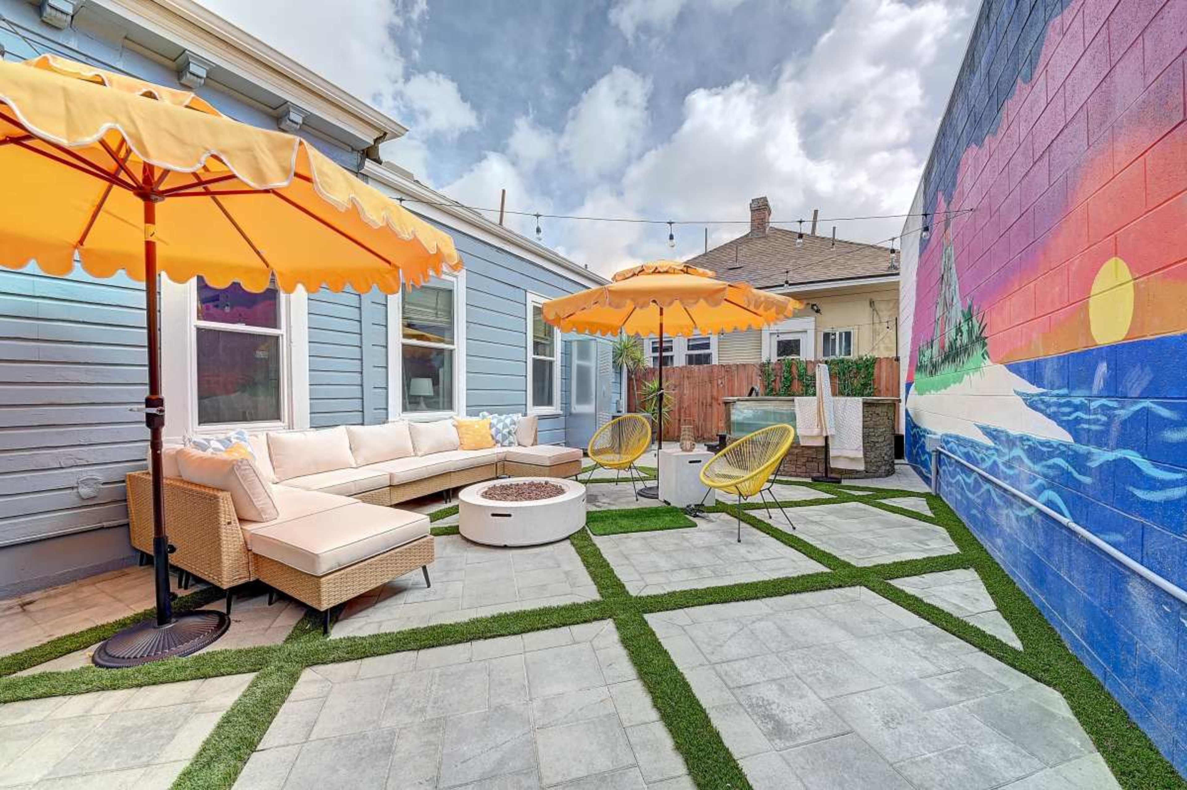 The image shows a patio area featuring a sectional couch, a fire pit, two yellow umbrellas, and a colorful mural on a wall.