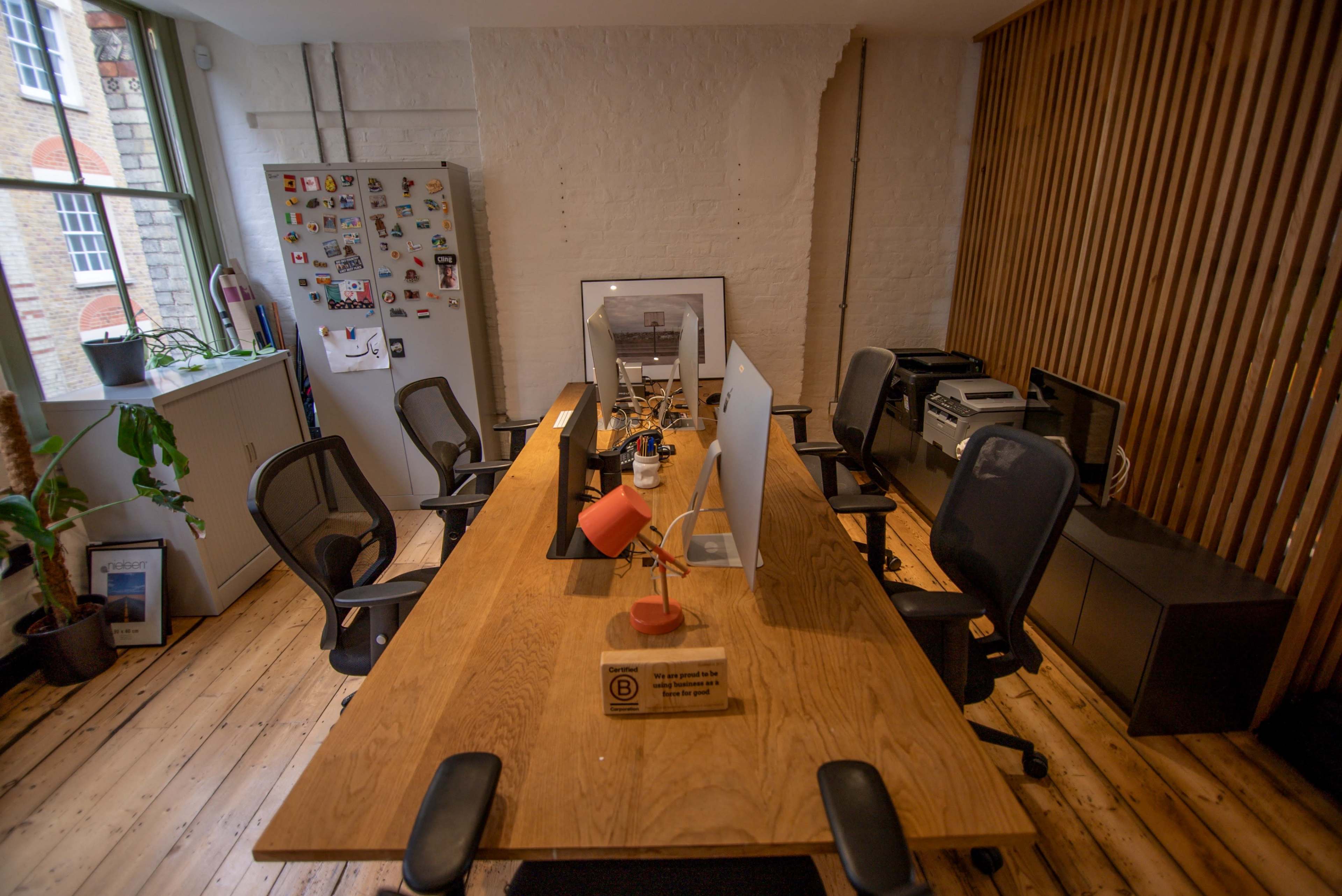 A spacious office features a long wooden table surrounded by black chairs, with a computer and lamp on the table, and a fridge covered in magnets in the background.