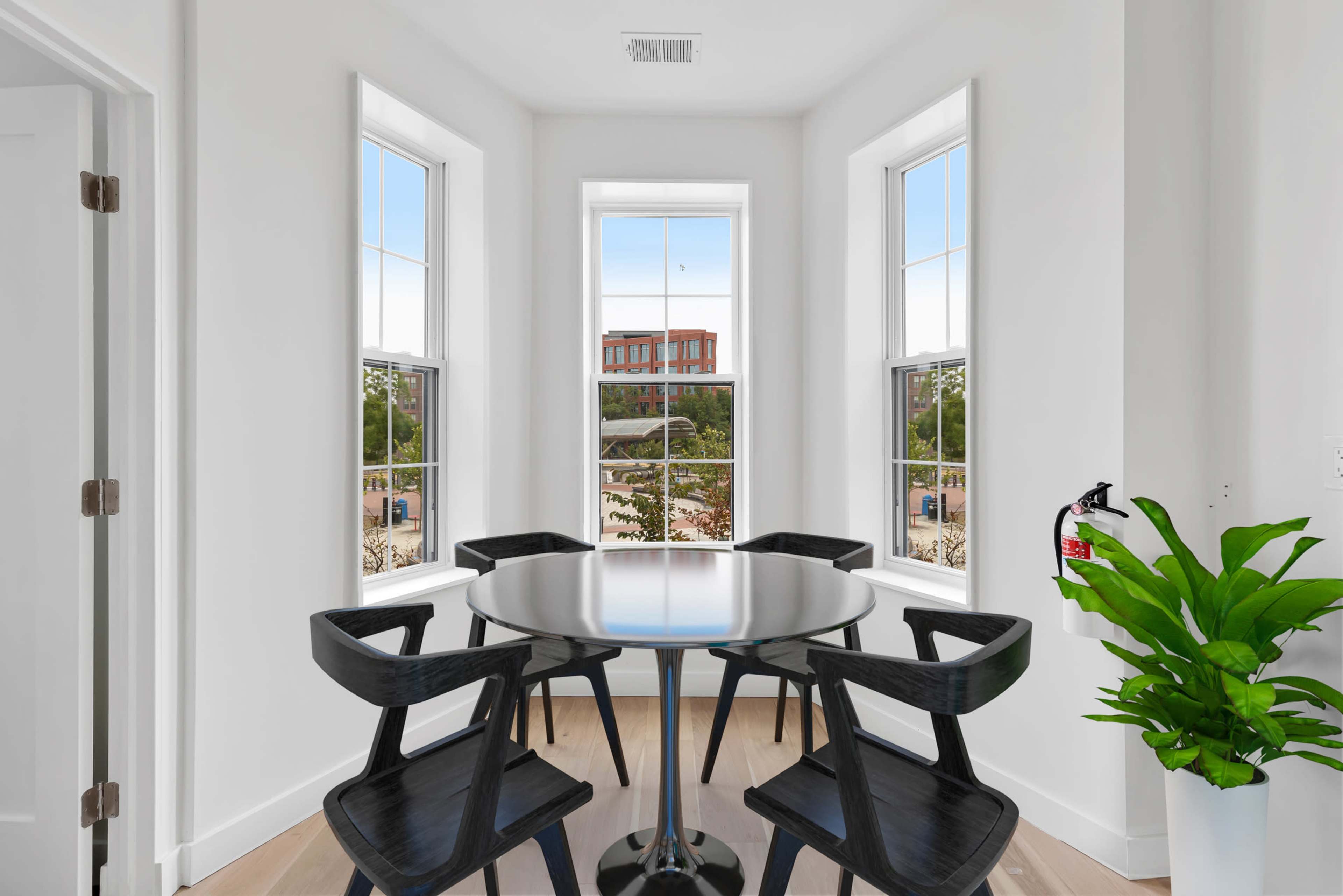 A bright, modern dining nook features a round black table surrounded by four black chairs, with large windows offering views of a landscaped area outside.