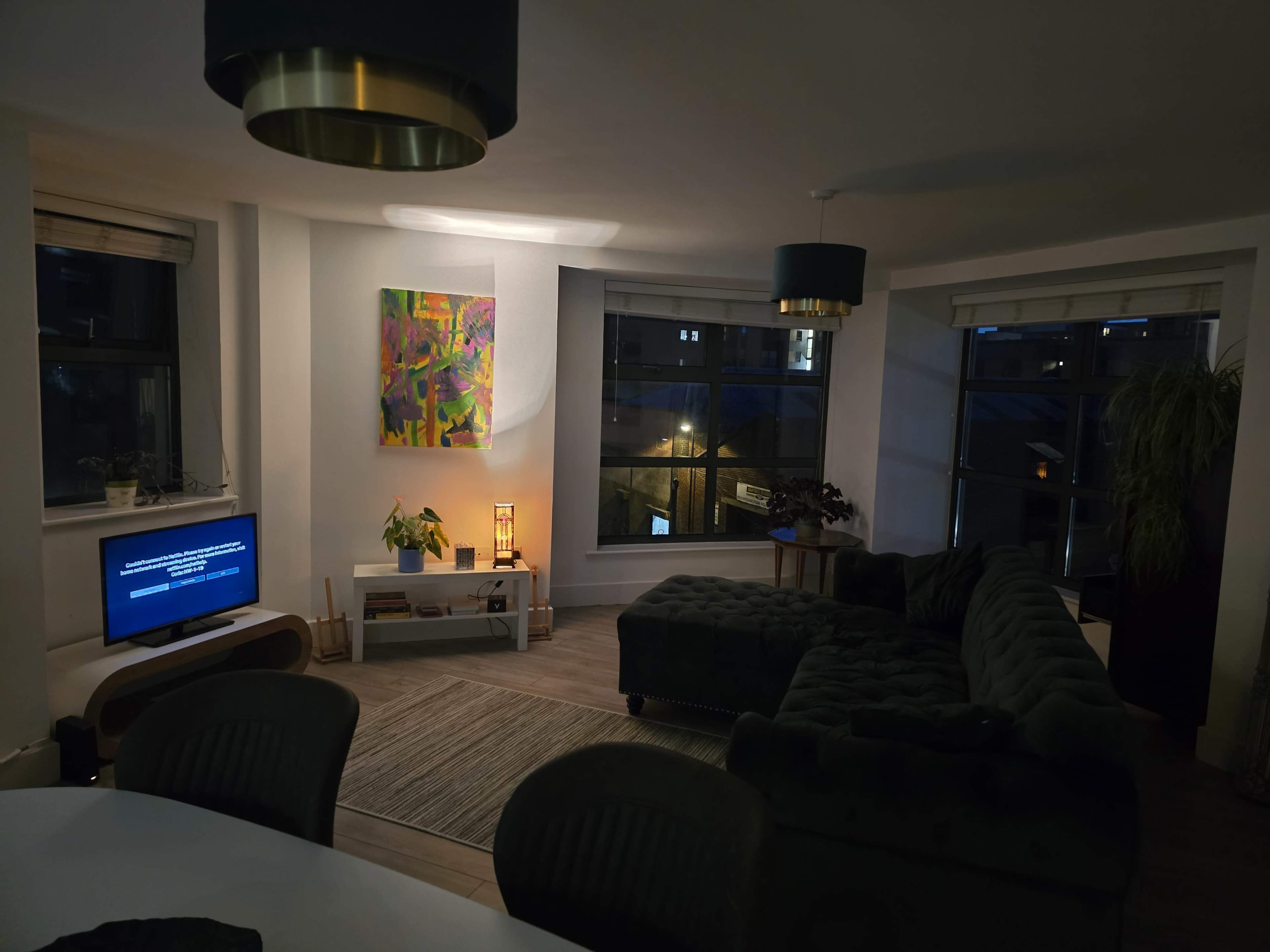 A modern living room features a sectional couch, a television on a white console, and large windows with city views.