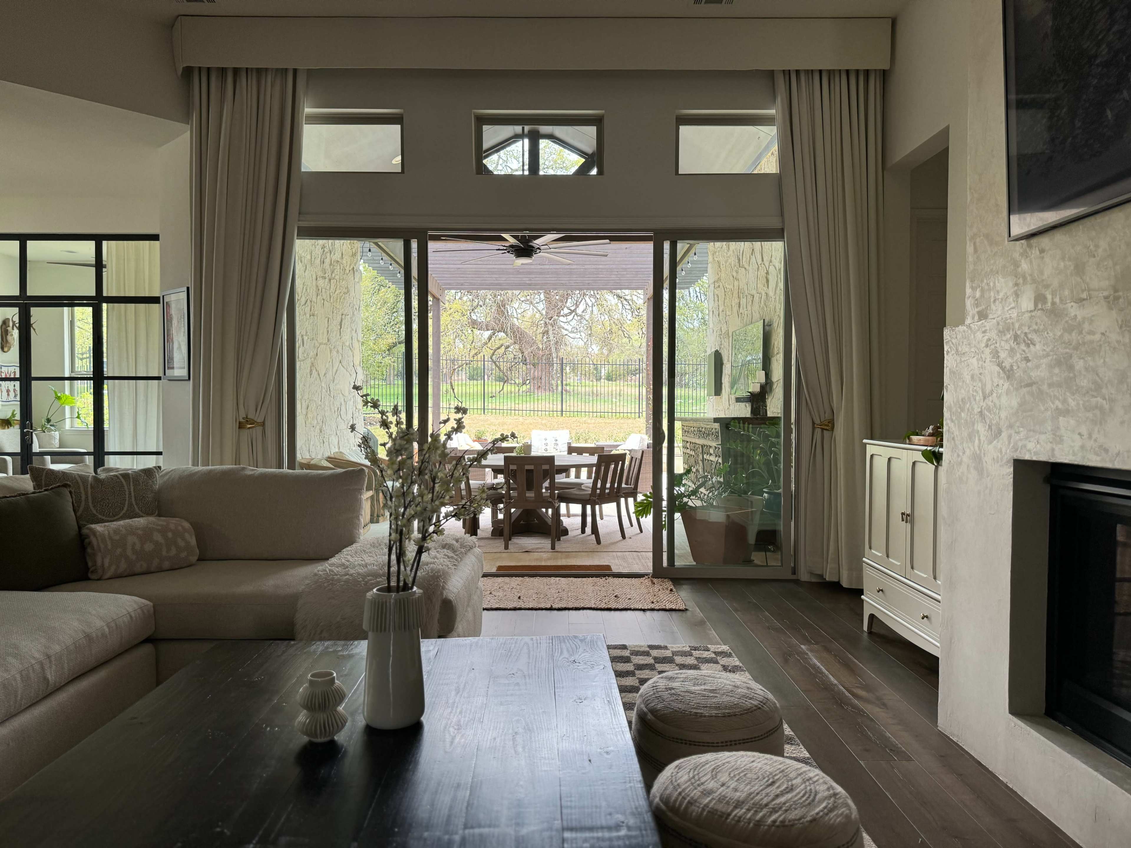 A living room with a large glass door leads to a patio area, featuring a dining table and chairs surrounded by greenery.