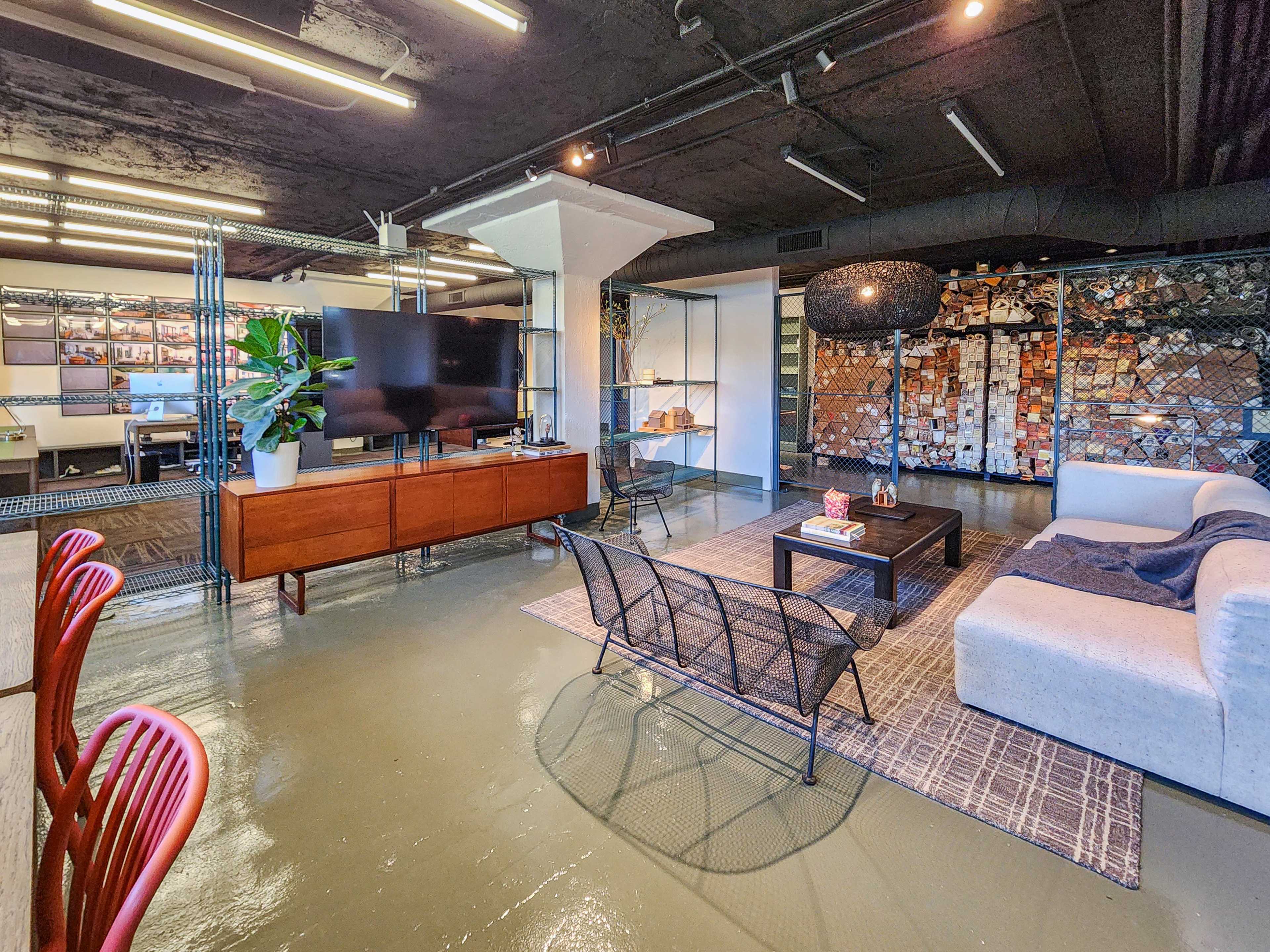 The image shows a modern office space featuring a seating area with a sofa and coffee table, a television, and a wall adorned with various images and materials, all set against a contemporary industrial backdrop.