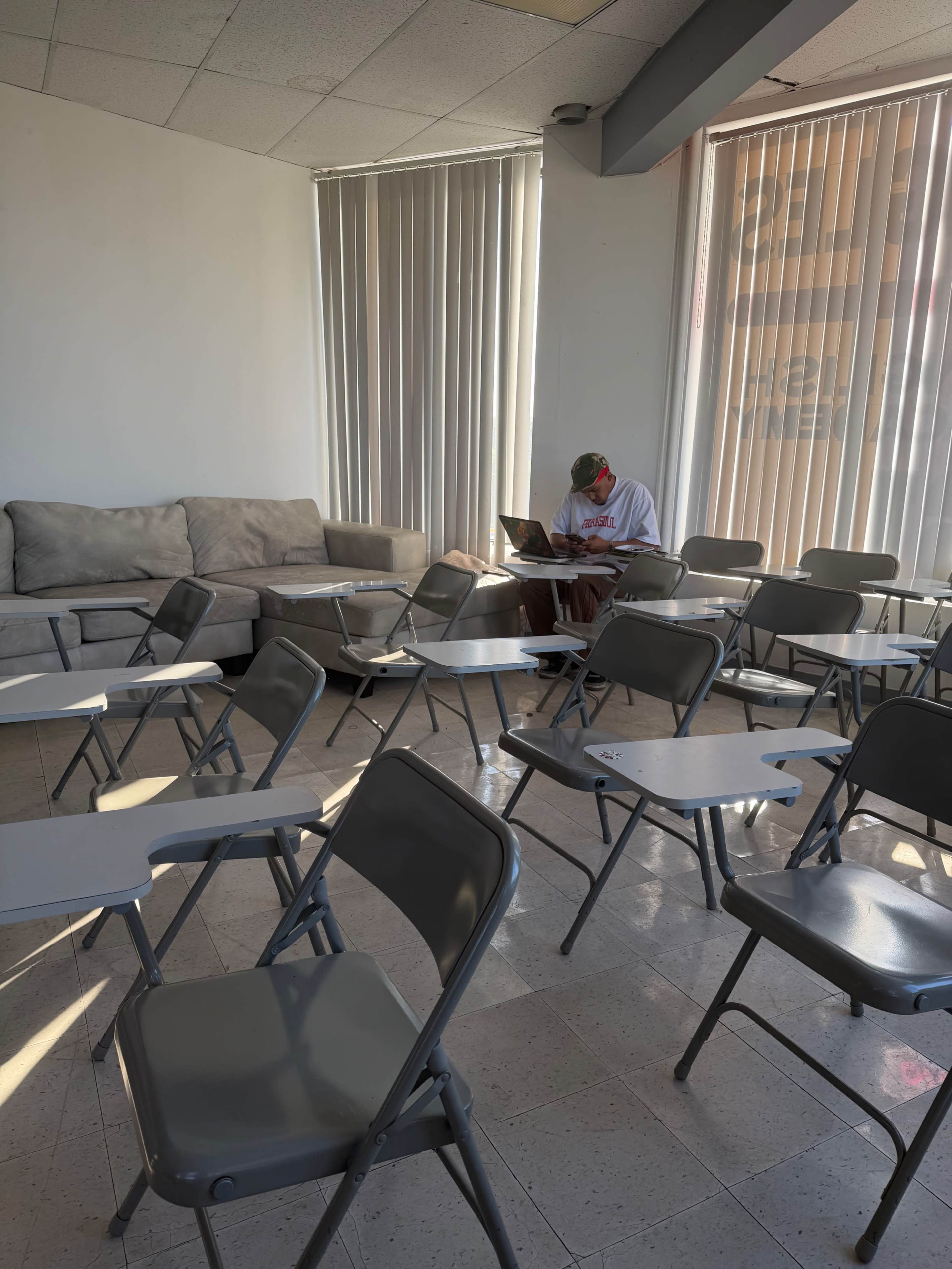 A student sits at a desk with a laptop in a nearly empty classroom, surrounded by rows of empty chairs and a couch against the wall.