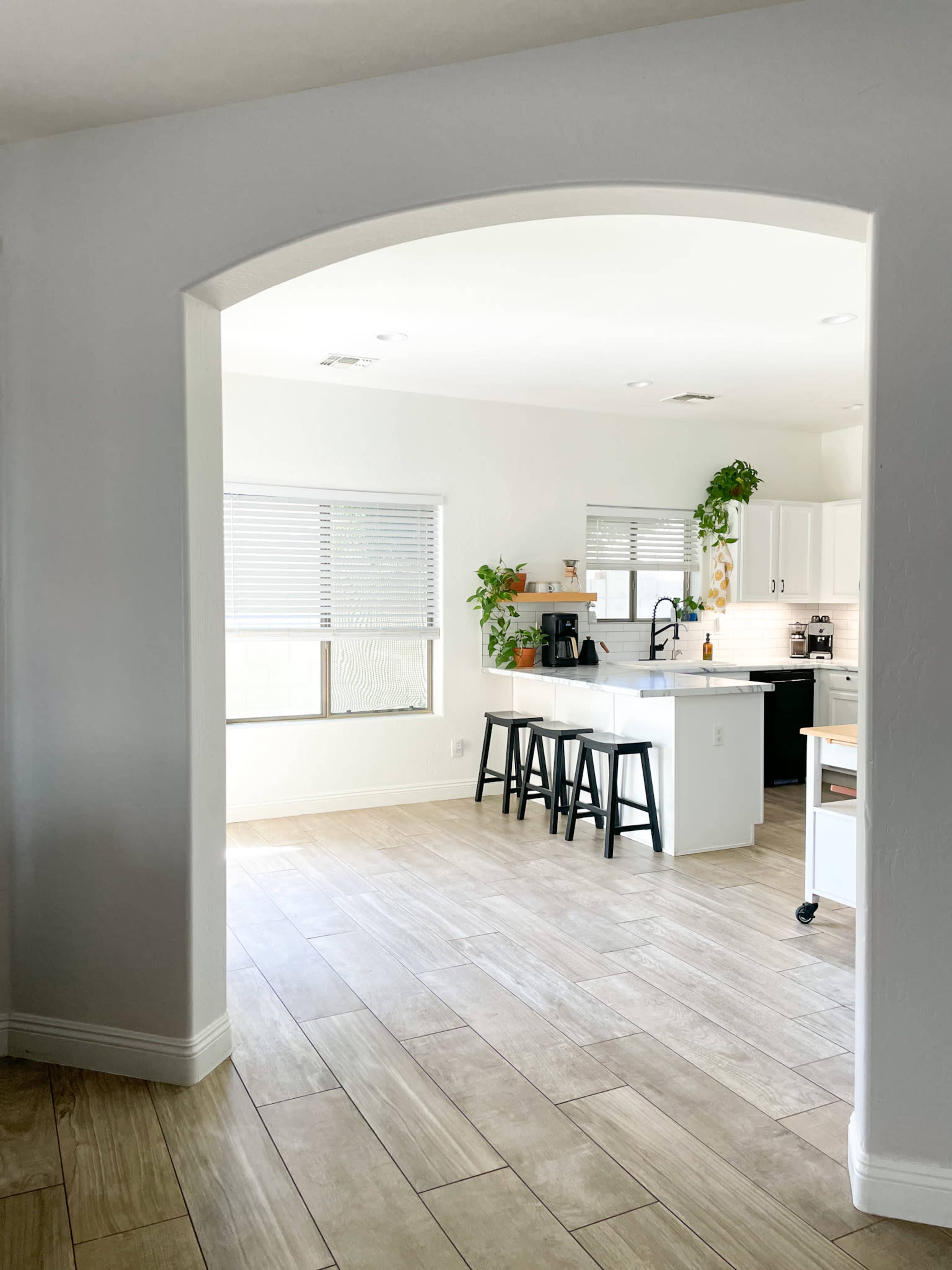 A bright kitchen features a white layout with a small table and black stools, seen through an arched doorway.