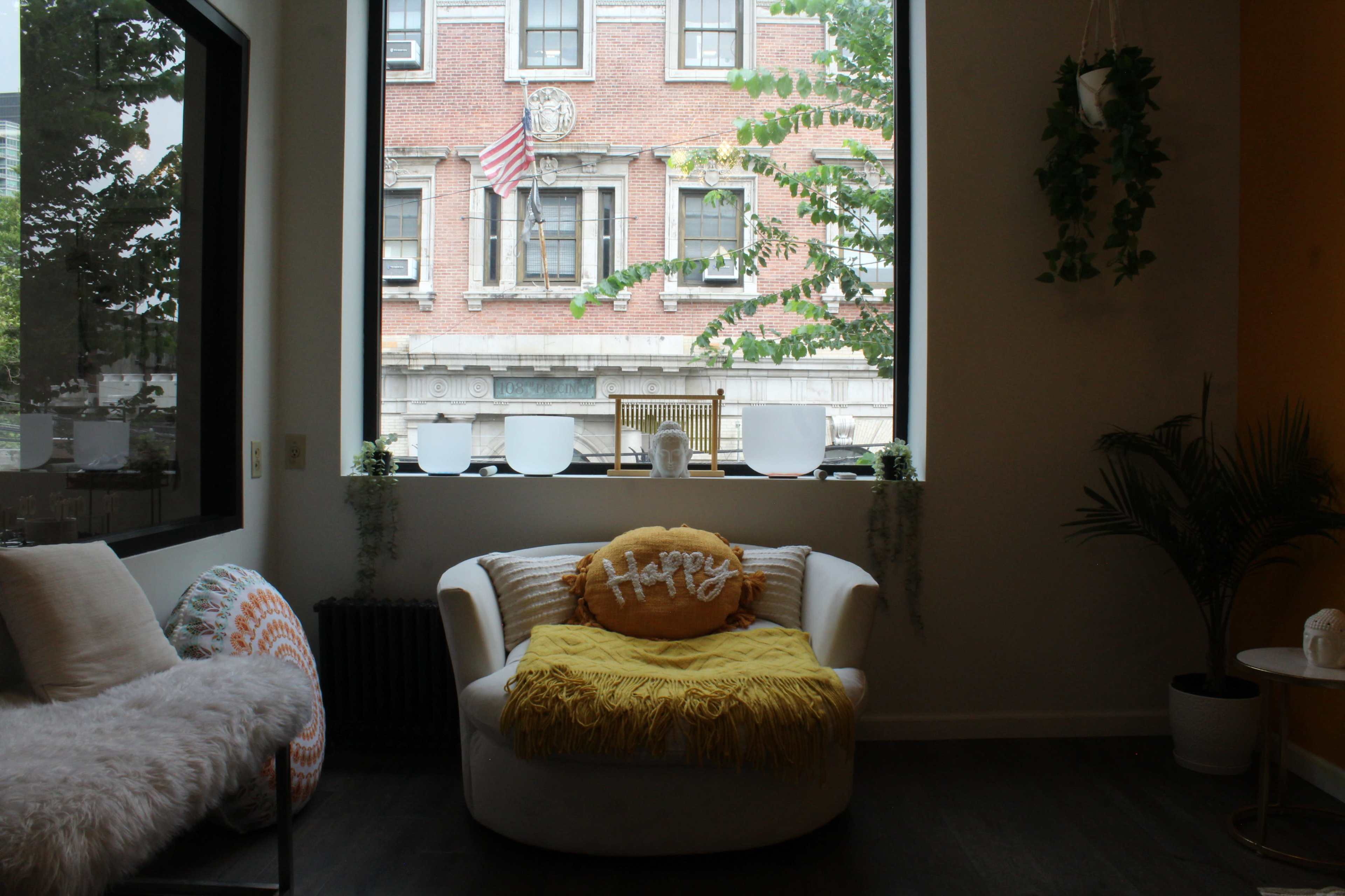 A cozy lounge area features a white armchair adorned with a yellow throw and a decorative pillow, positioned in front of a large window that offers a view of a brick building and an American flag.