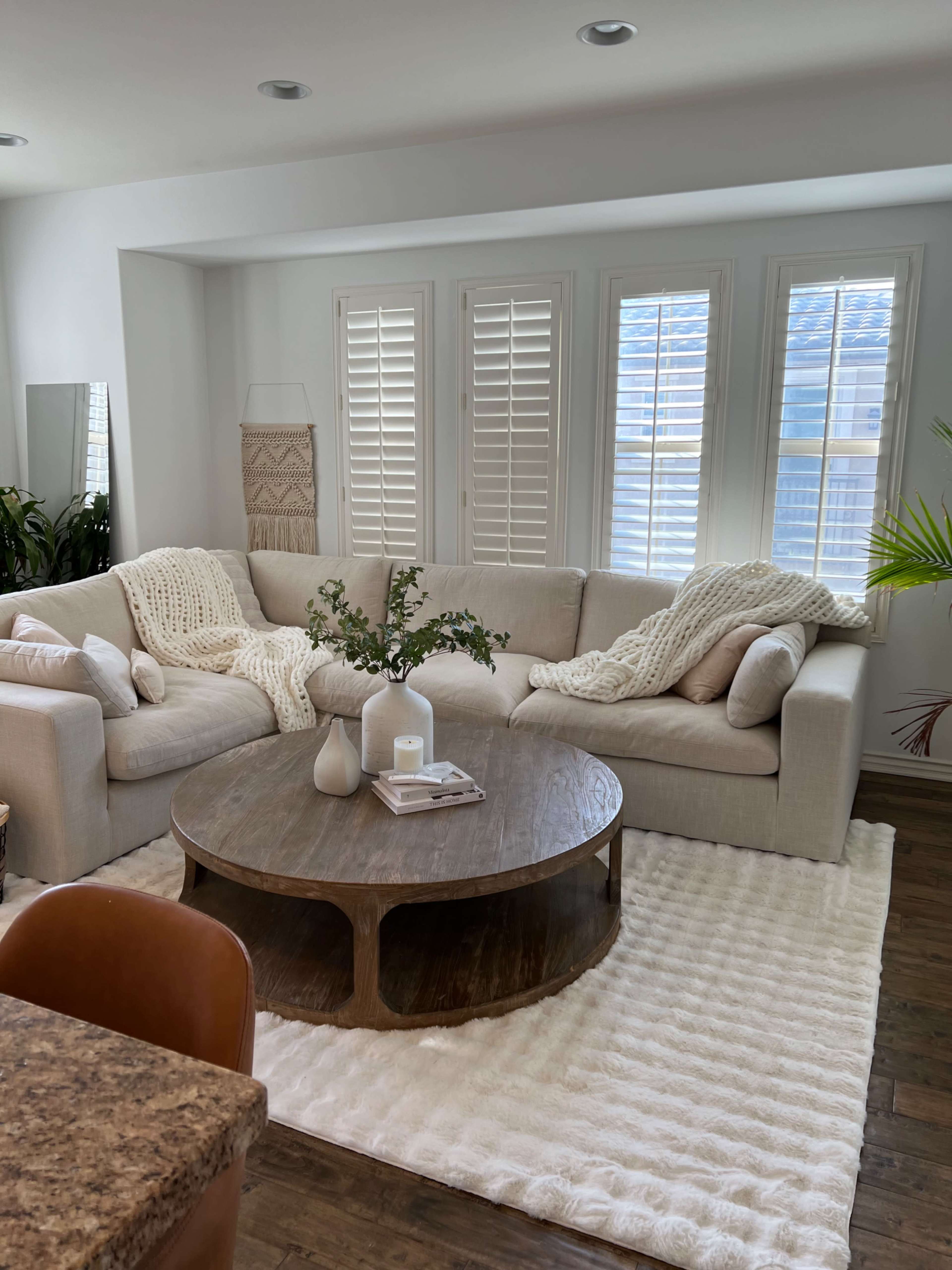The image shows a light-colored living room featuring a sectional sofa with a textured throw, a round wooden coffee table, and large windows with white shutters.