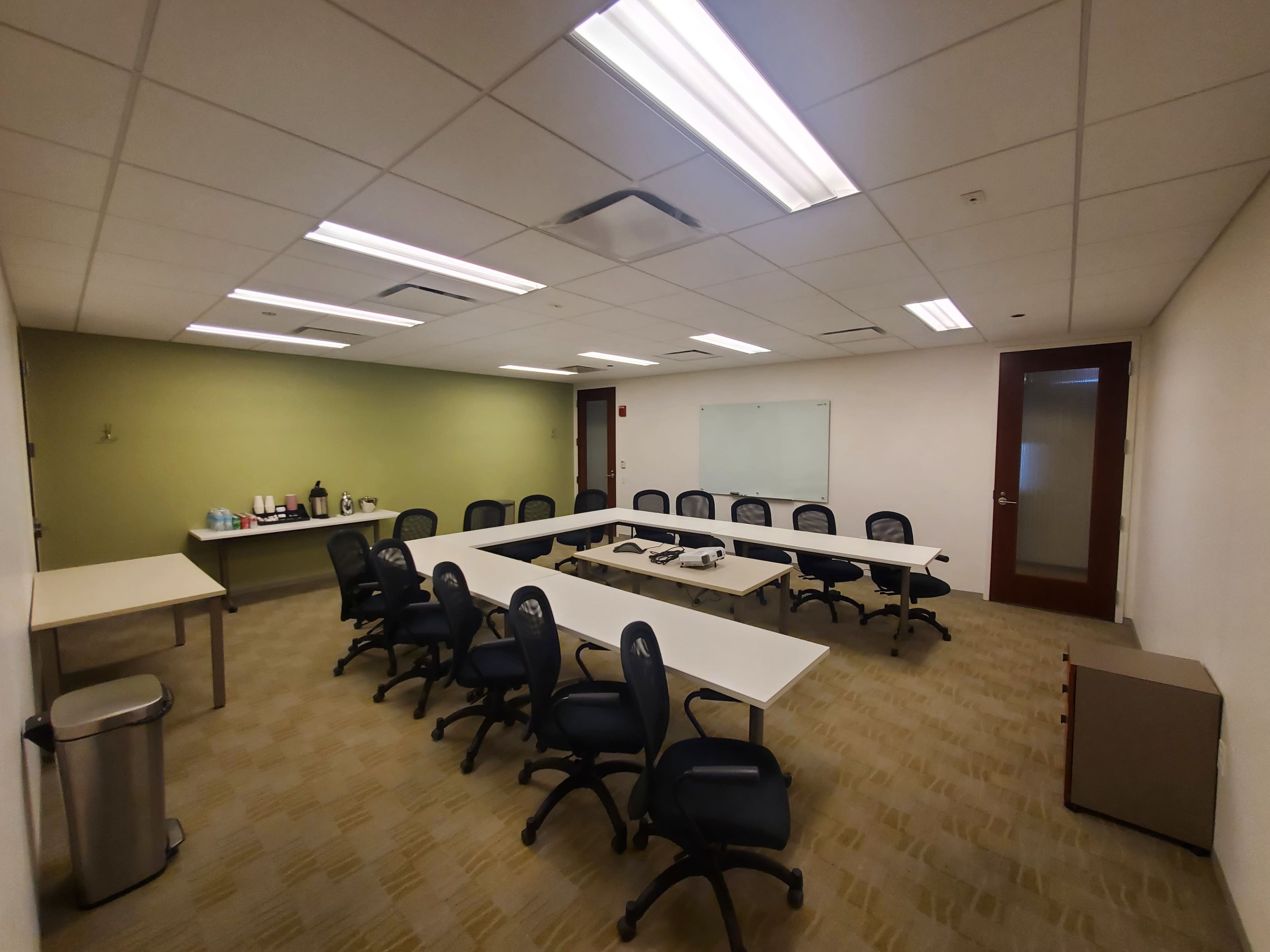 A conference room features a long, white table surrounded by black chairs, with a service area containing beverages and a whiteboard.