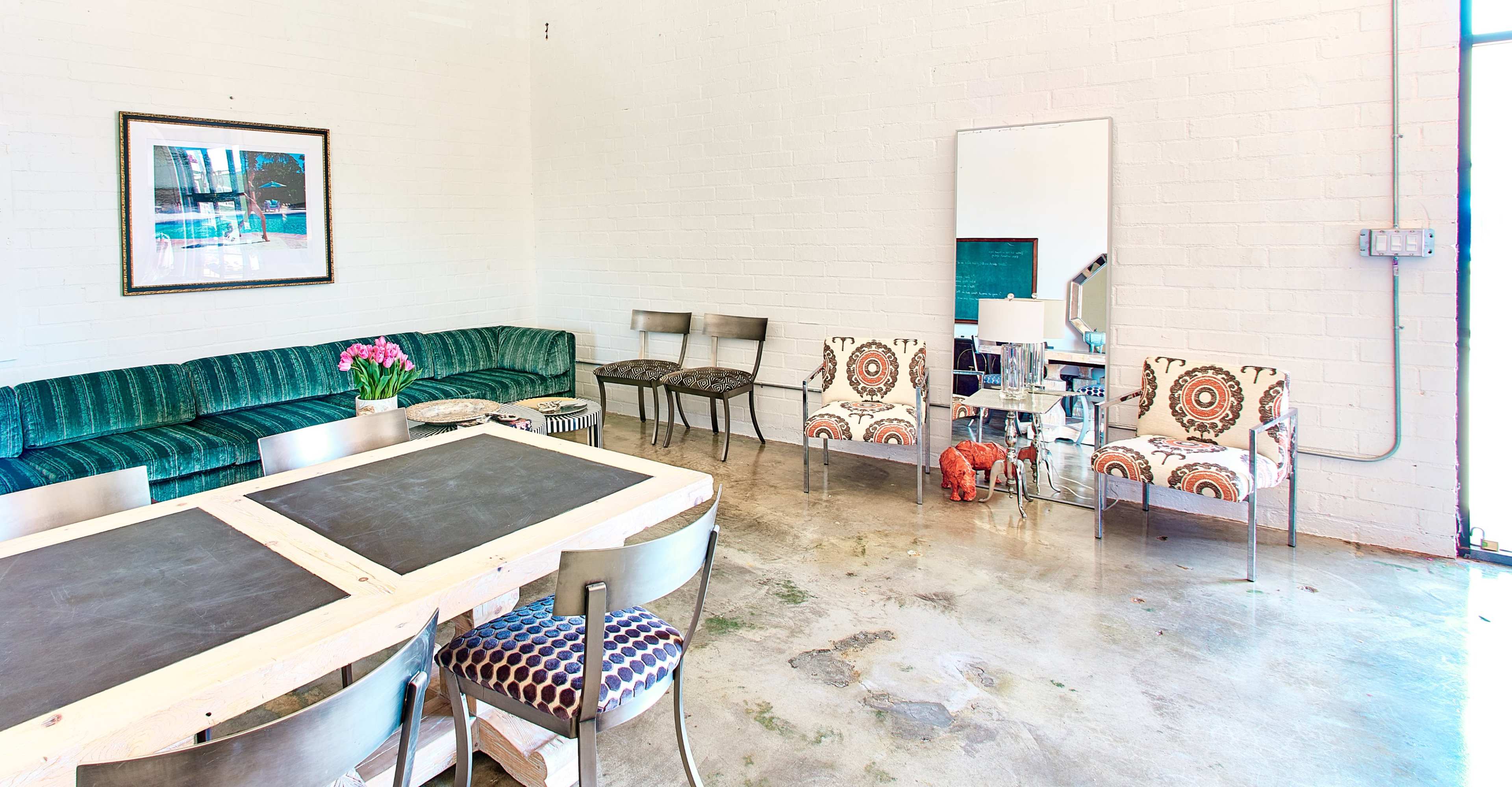 The image shows a spacious room with a green velvet sofa, a dining table with black surfaces, and various seating options, including patterned chairs, next to a large mirror and an abstract painting.