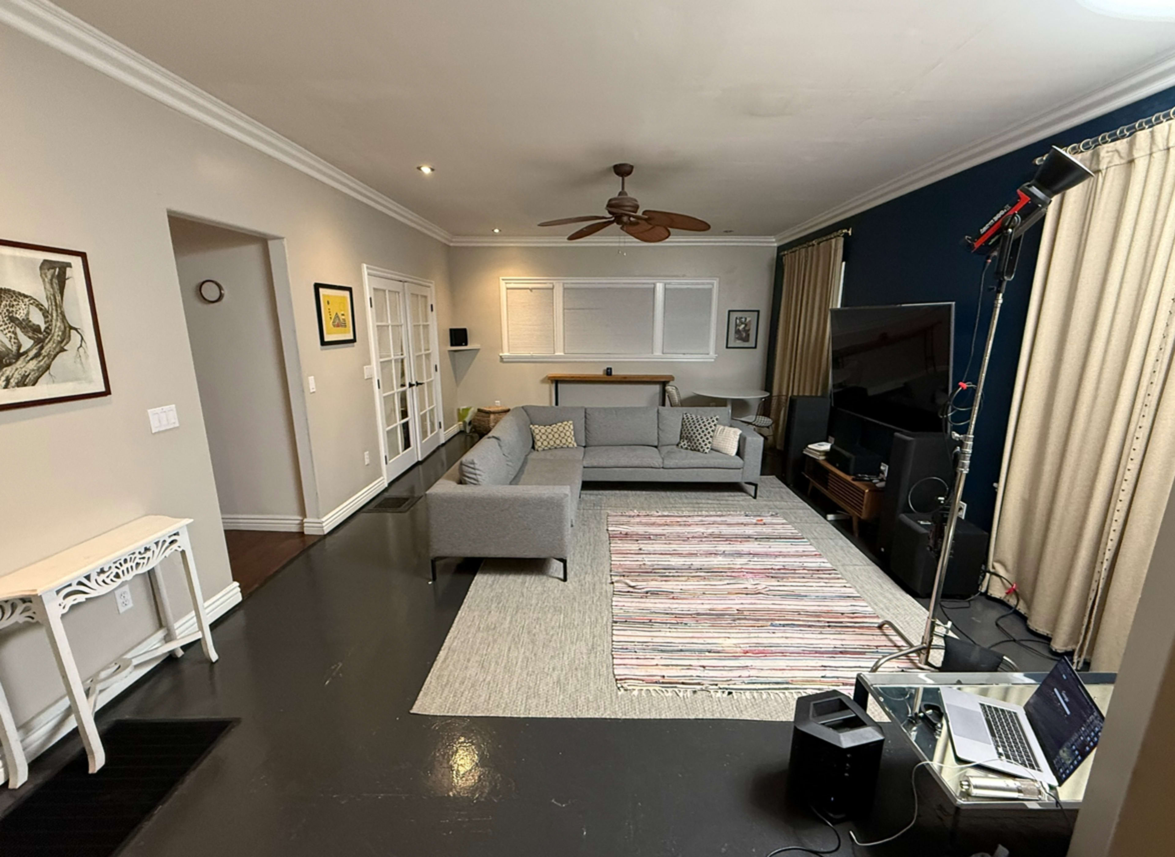 The image shows a living room with a gray sofa, a light rug, a wooden coffee table, and a television setup in the corner, illuminated by studio lighting.