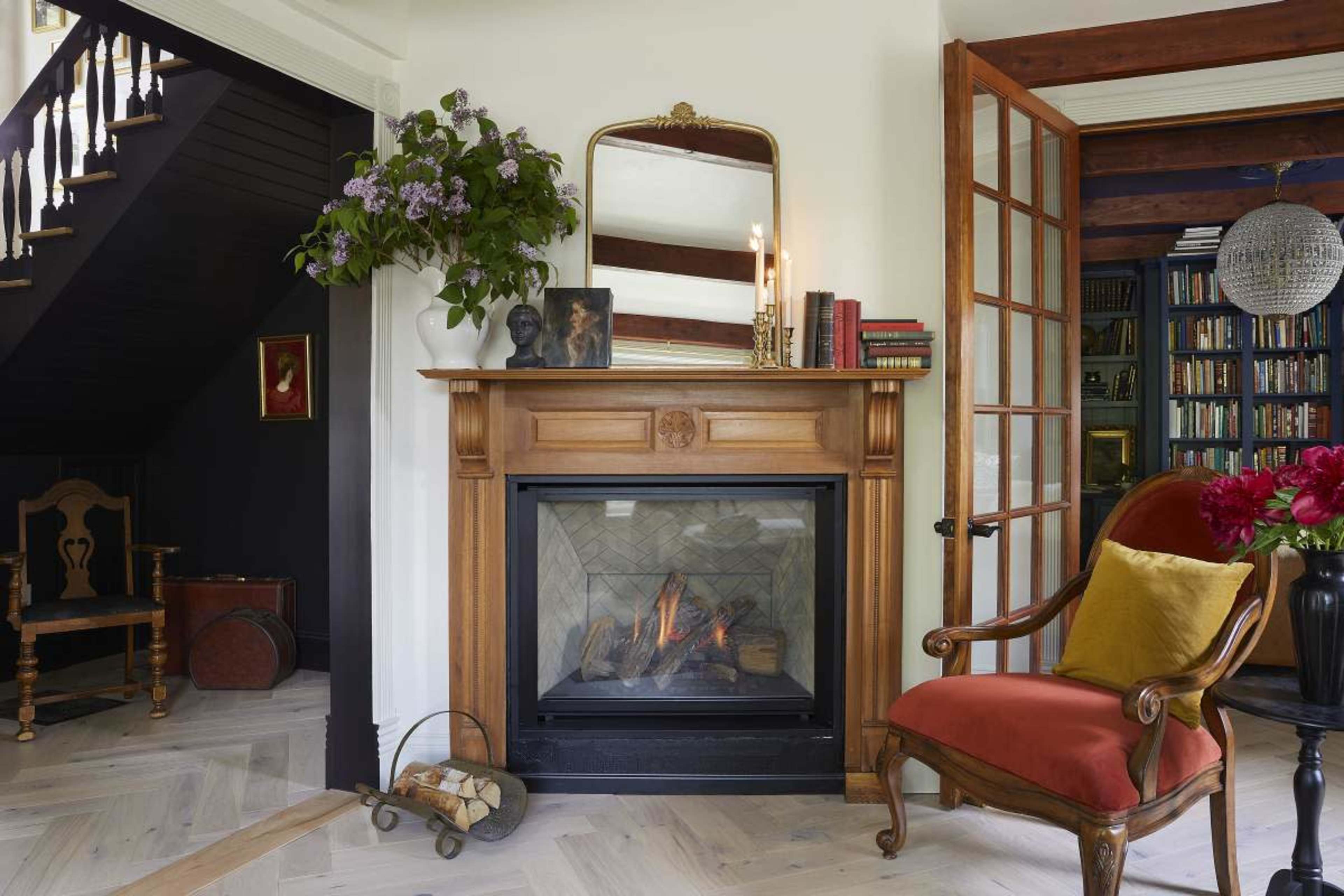 A modern living room features a wooden fireplace with a mirror above it, a cozy armchair, and bookshelves in the background.