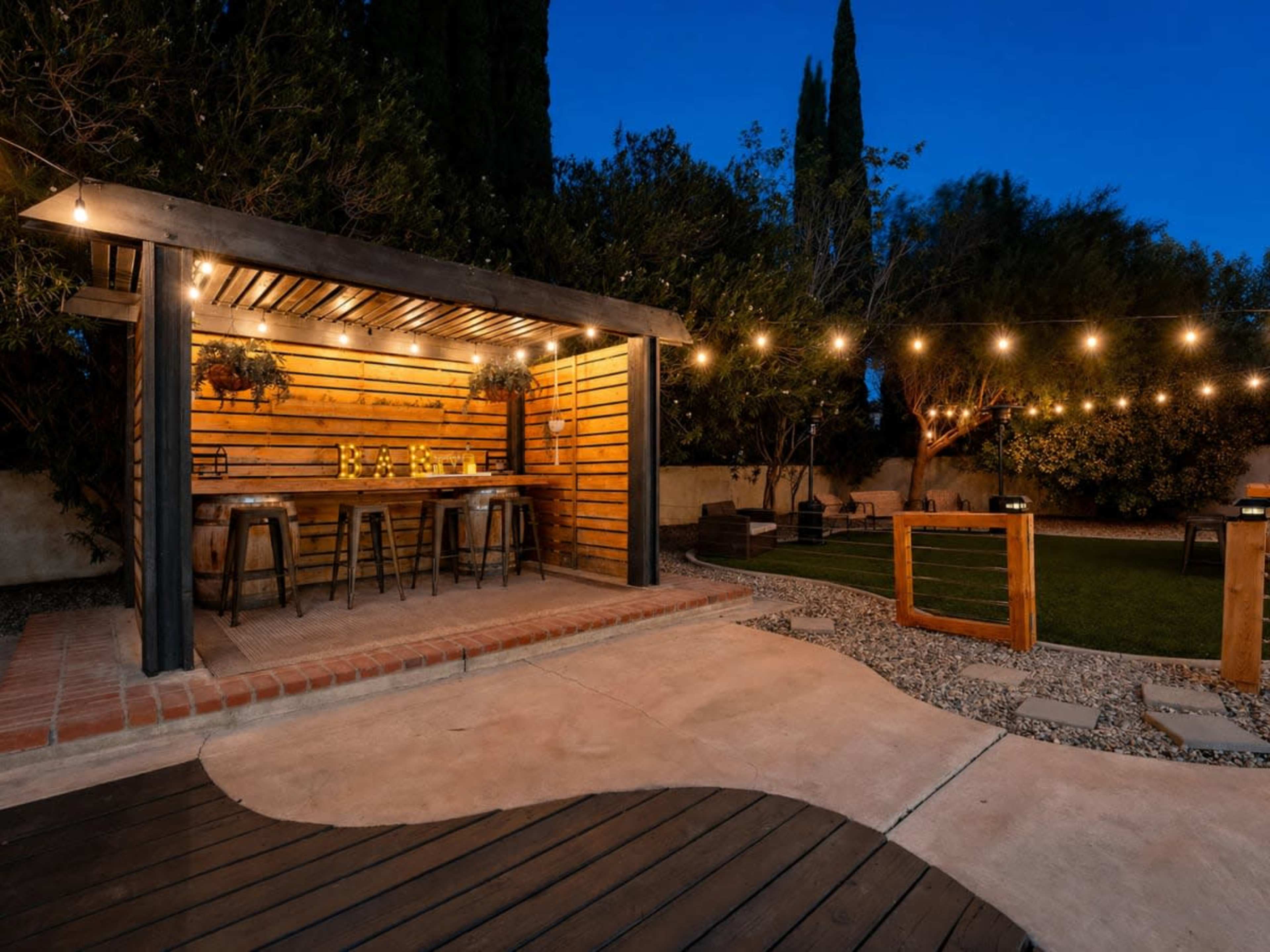 The image shows a backyard area featuring a wooden bar with stools, string lights overhead, and a lawn space surrounded by trees.