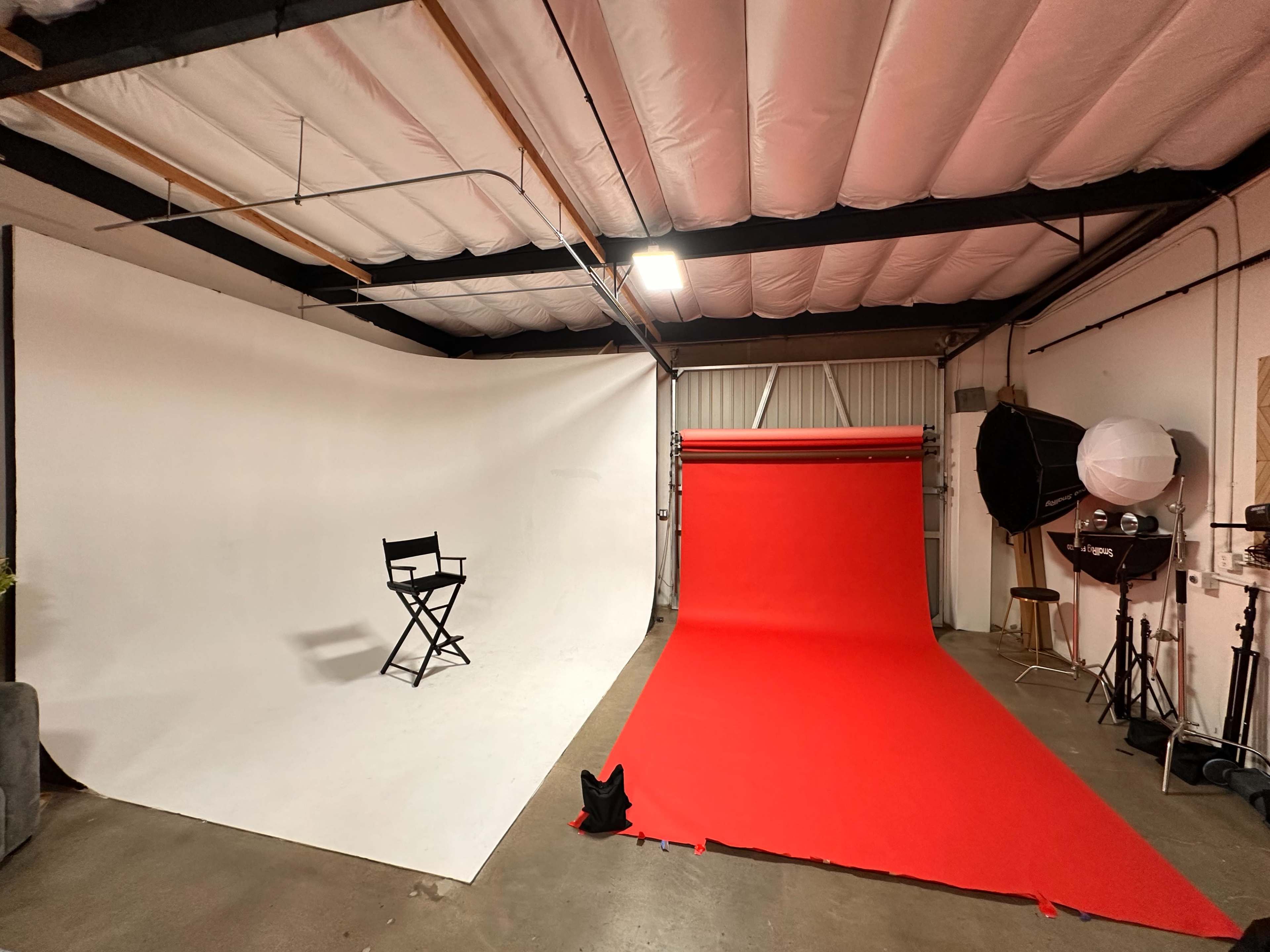 The image shows a photography studio with a white backdrop on one side, a red backdrop on the other, and a director's chair positioned in front of the white wall.