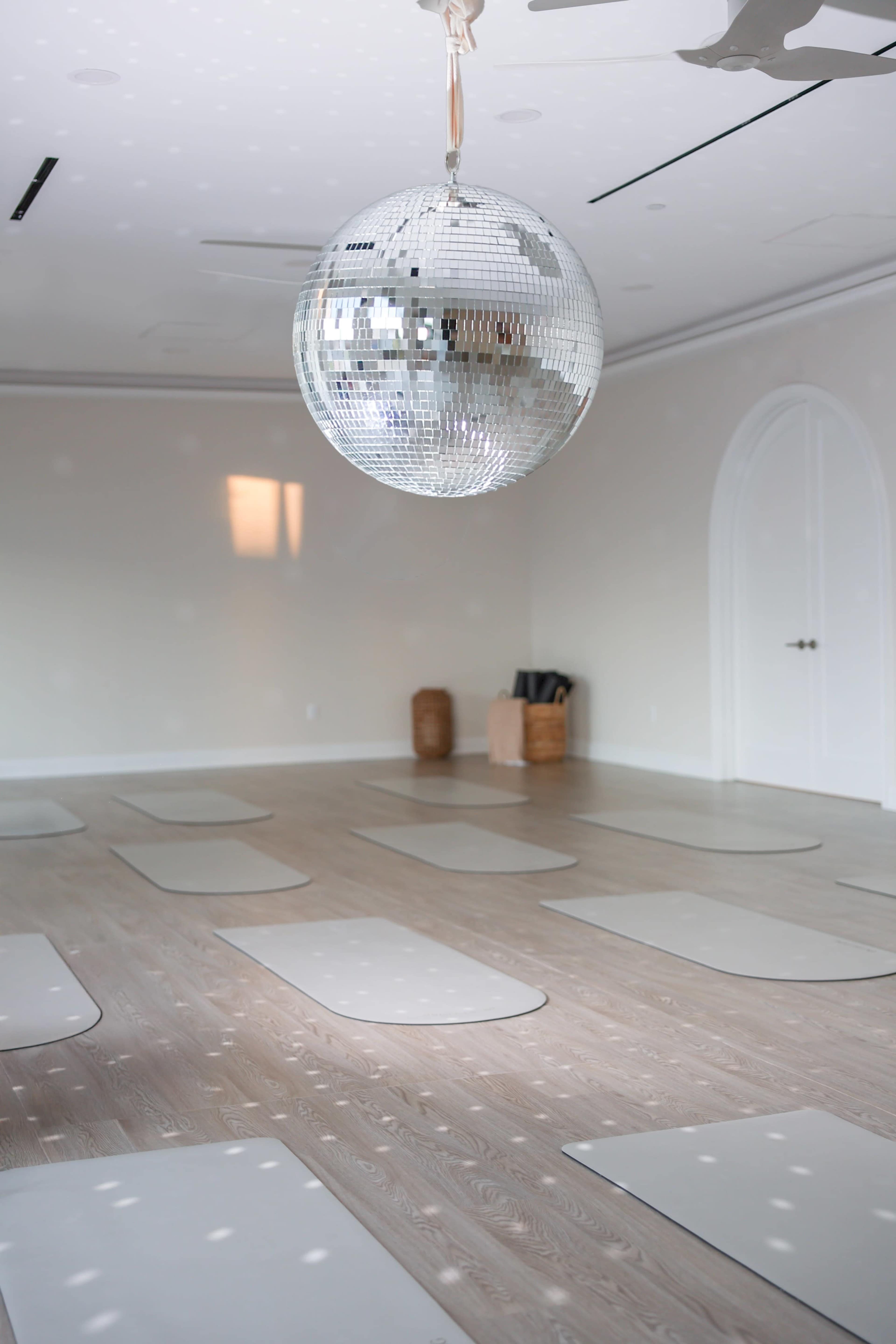 A mirrored disco ball hangs from the ceiling above a spacious yoga studio with several mats arranged on the wooden floor.