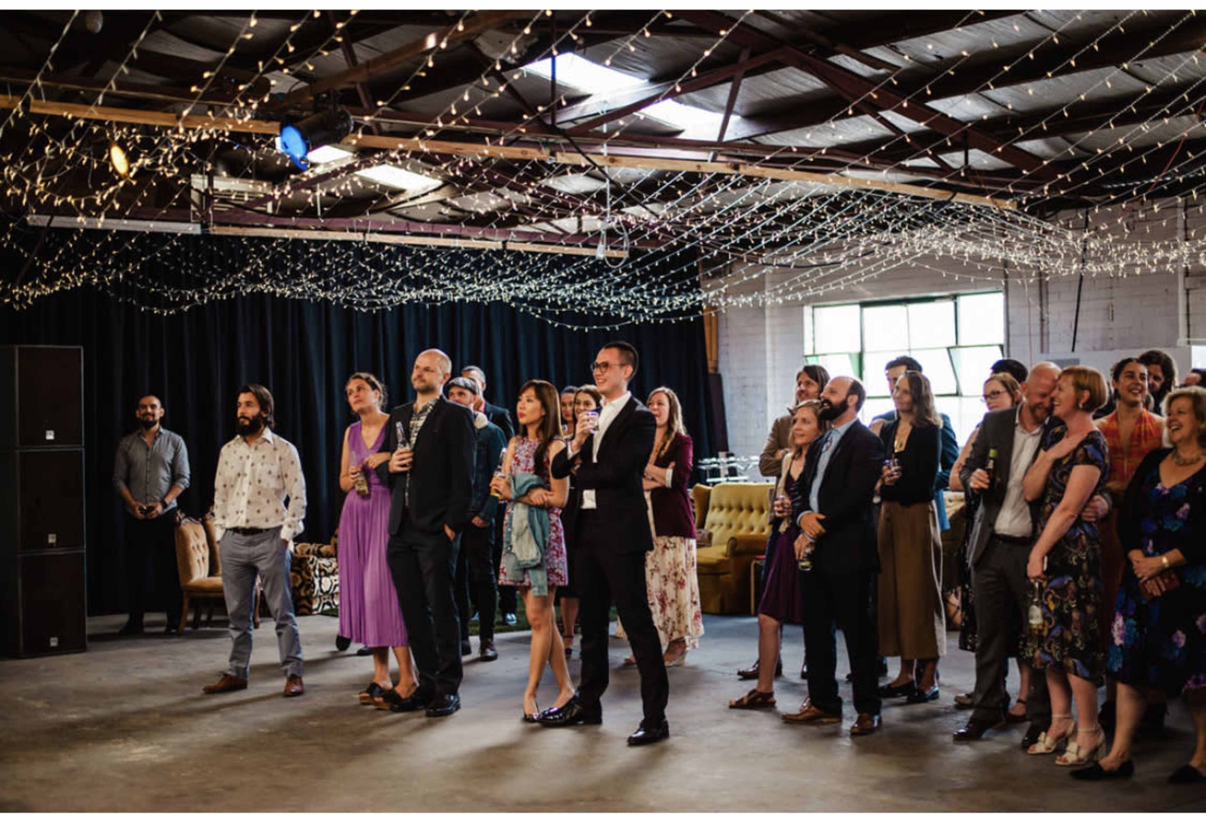 A group of people in formal and semi-formal attire are gathered in a decorated venue with string lights overhead, watching a presentation or performance.