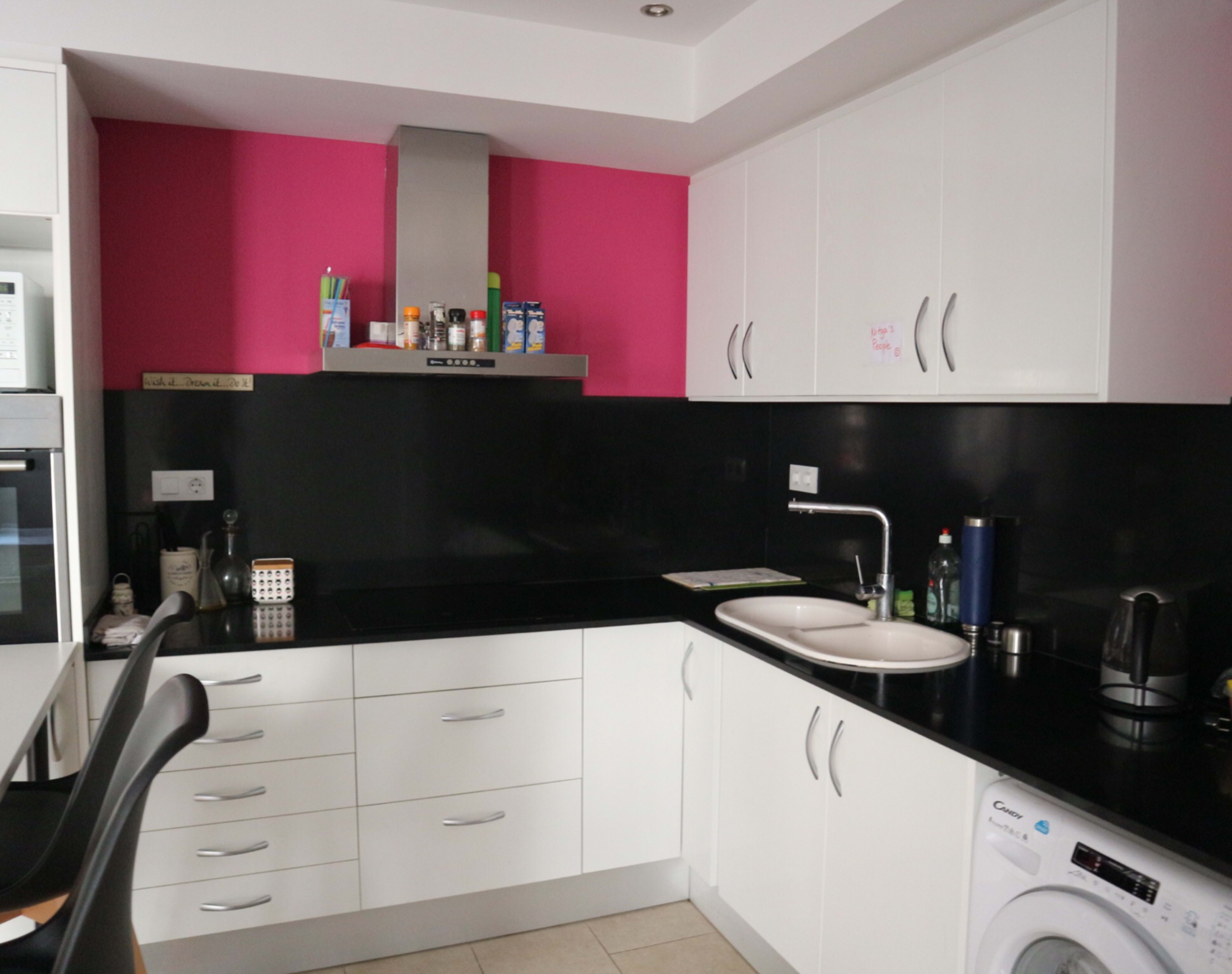 A modern kitchen features black countertops, white cabinetry, a pink accent wall, and stainless steel appliances.