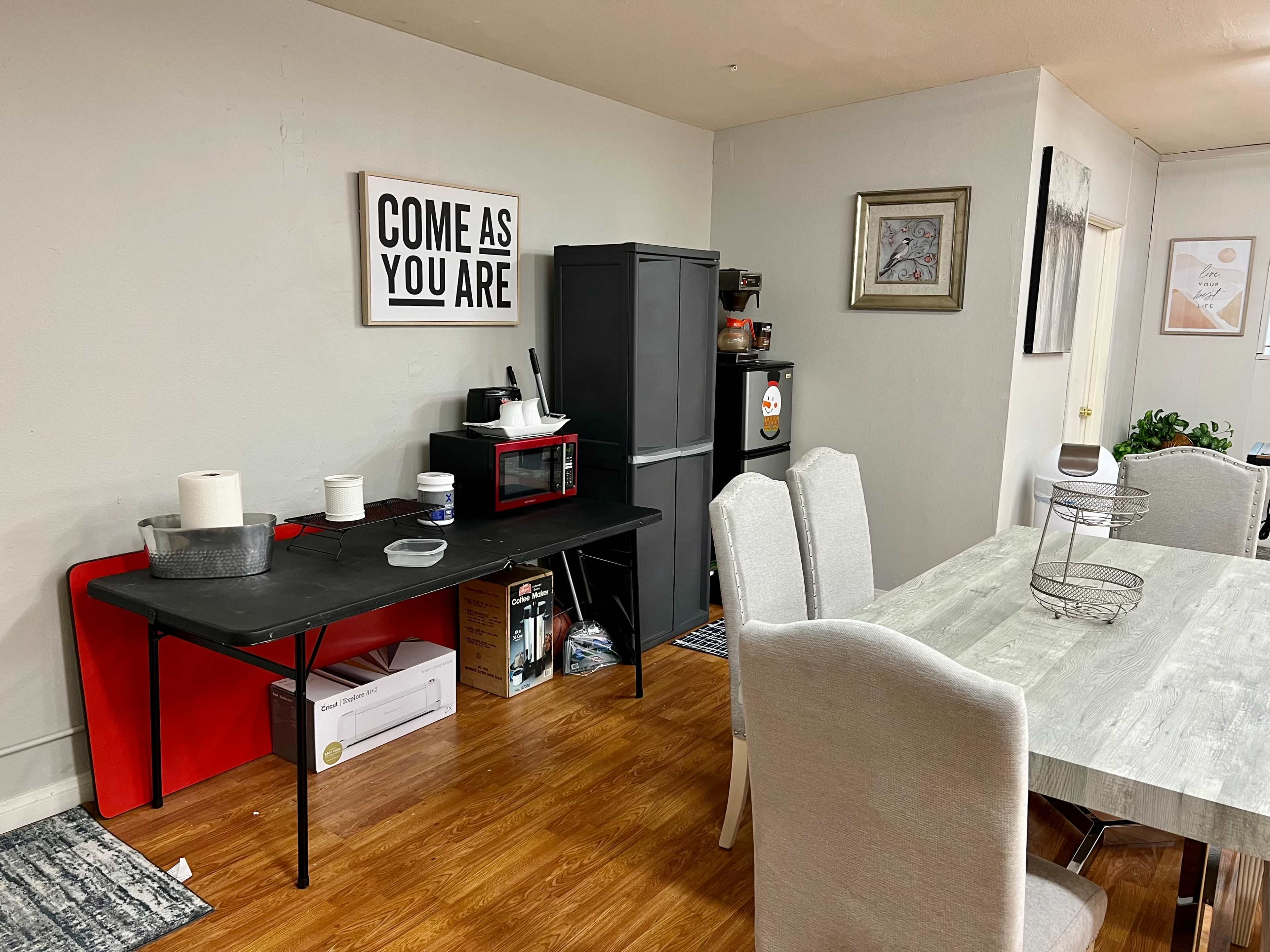A dining area with a gray table and chairs, a black table with kitchen items, and a small kitchen appliance setup against a wall featuring a large framed sign that reads "COME AS YOU ARE."