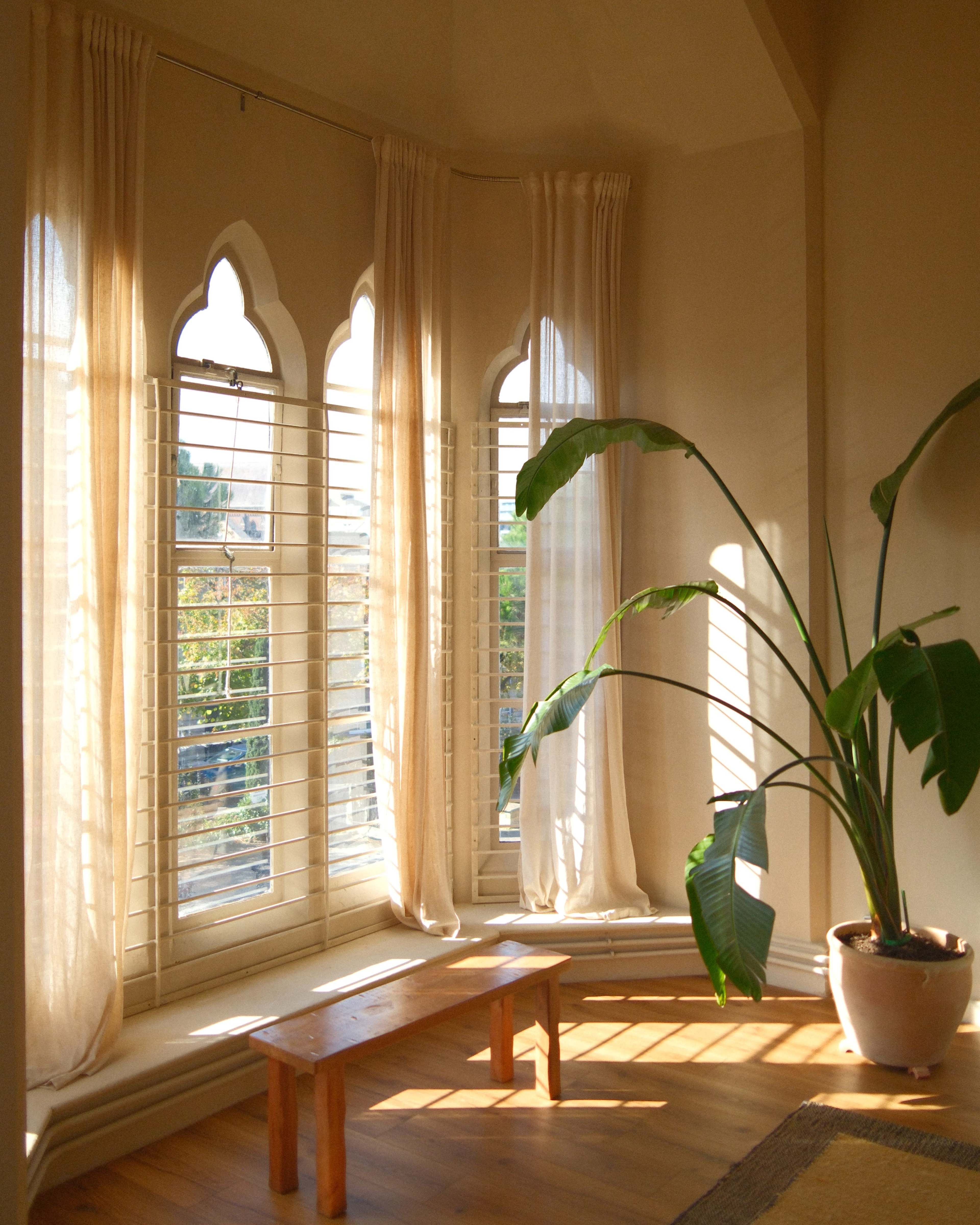 The image shows a sunlit bay window with decorative curtains, a wooden bench, and a large potted plant.