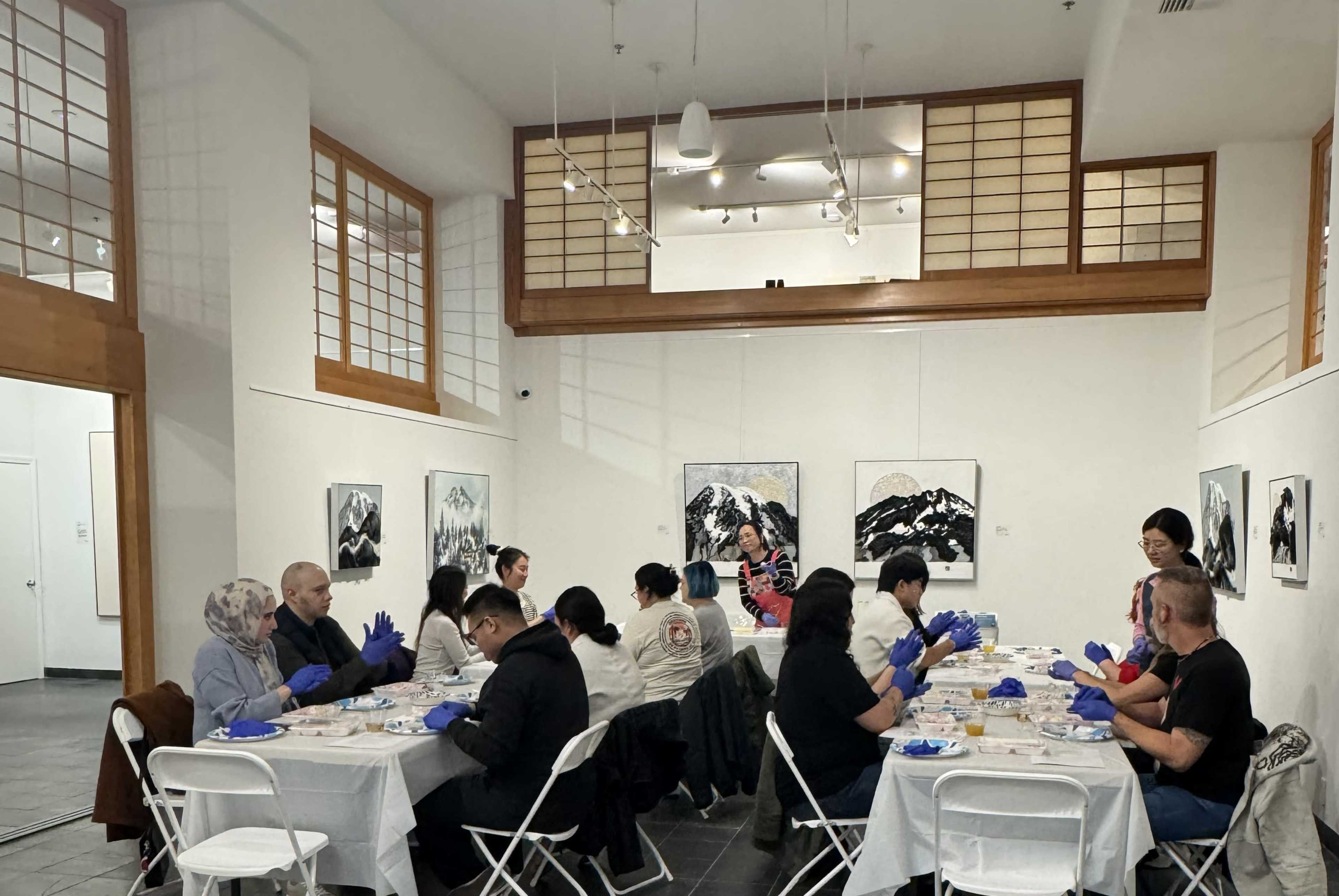 A group of people wearing blue gloves sits at tables in an art gallery, engaging in an activity while surrounded by paintings on the walls.