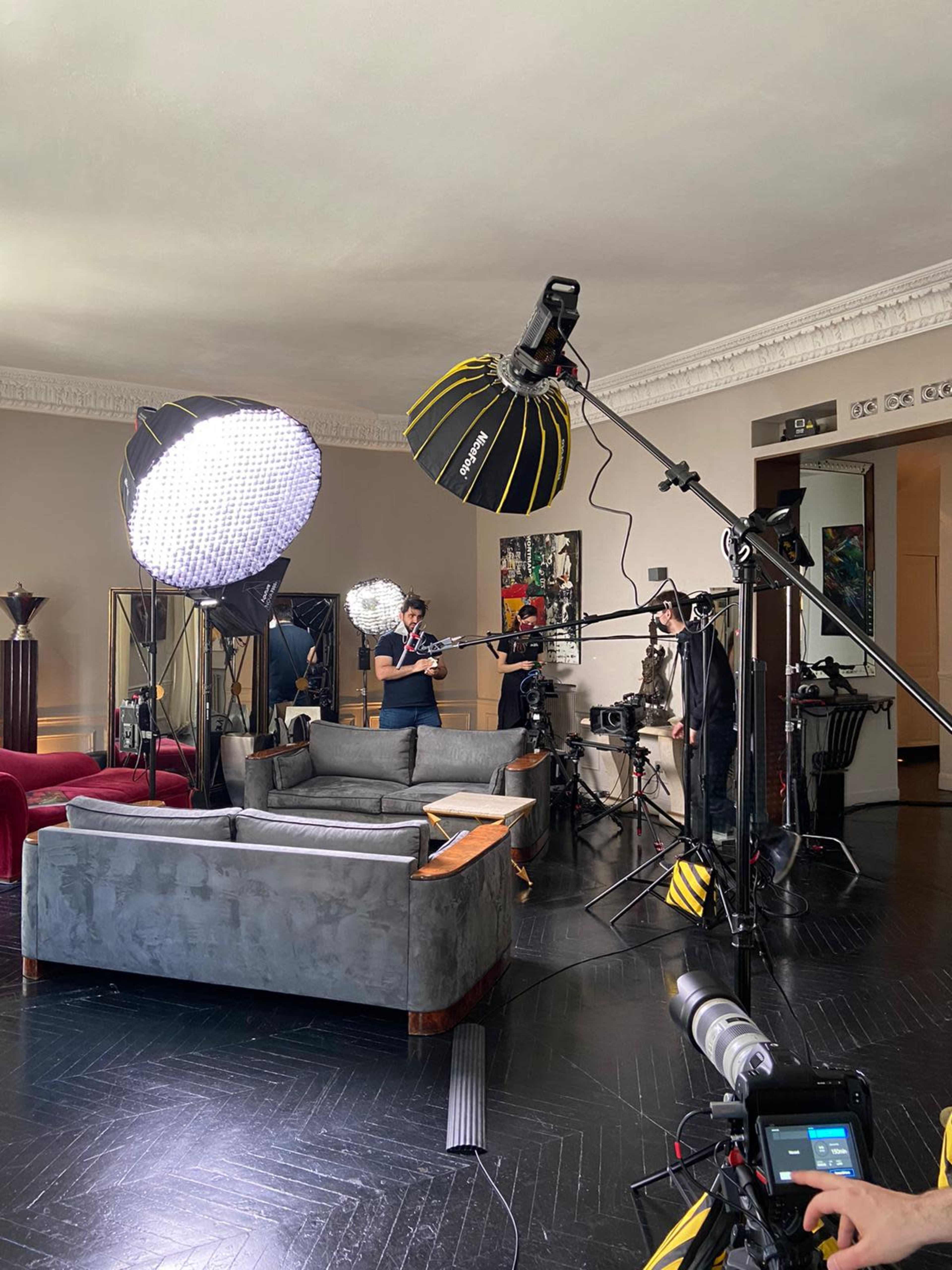 A film crew is setting up equipment in a well-lit living room, featuring a large gray sofa and various lighting tools.