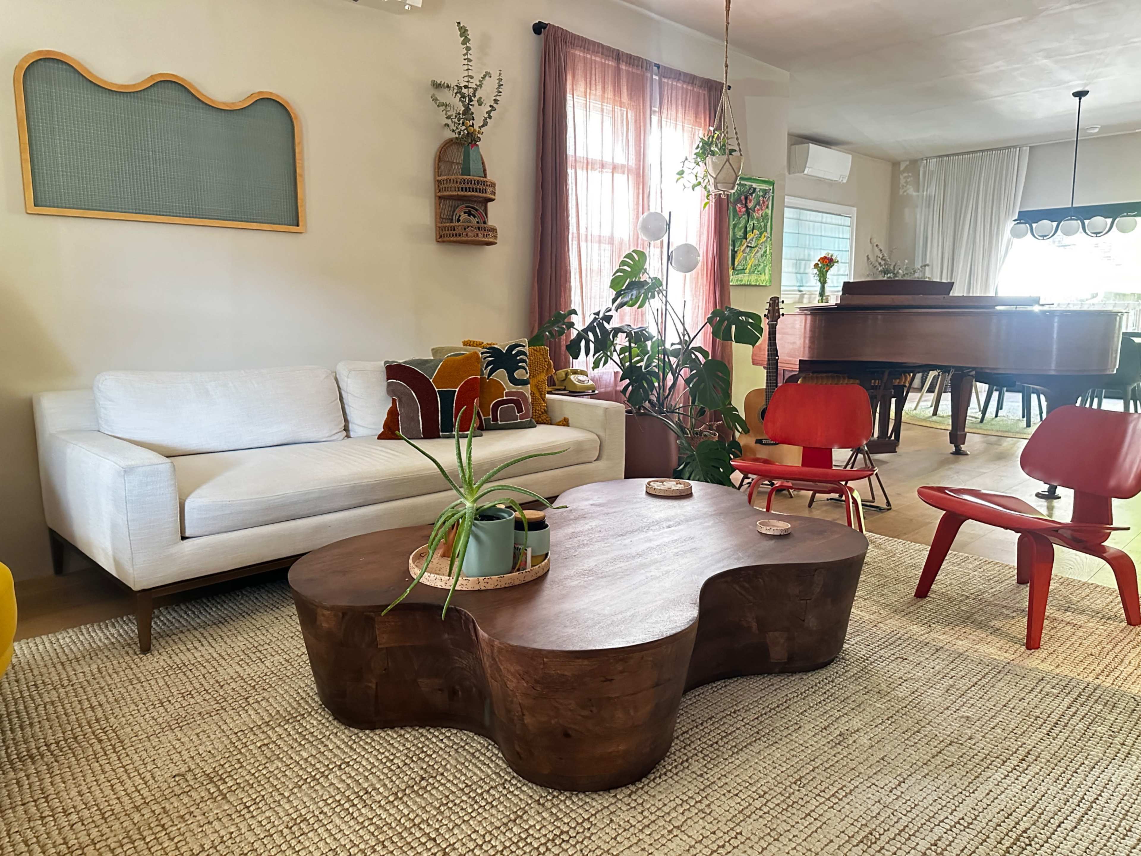 The living room features a white sofa, a unique wooden coffee table, red chairs, and various plants, with a piano in the background.