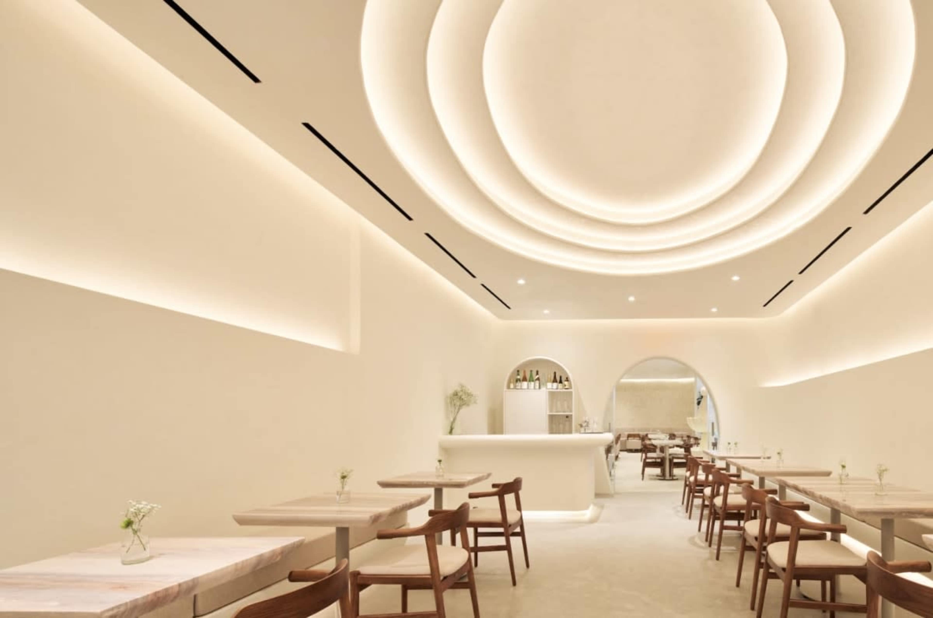 The image shows a modern restaurant with light-colored walls, minimalistic wooden furniture, and a uniquely designed ceiling that features circular recessed lighting.