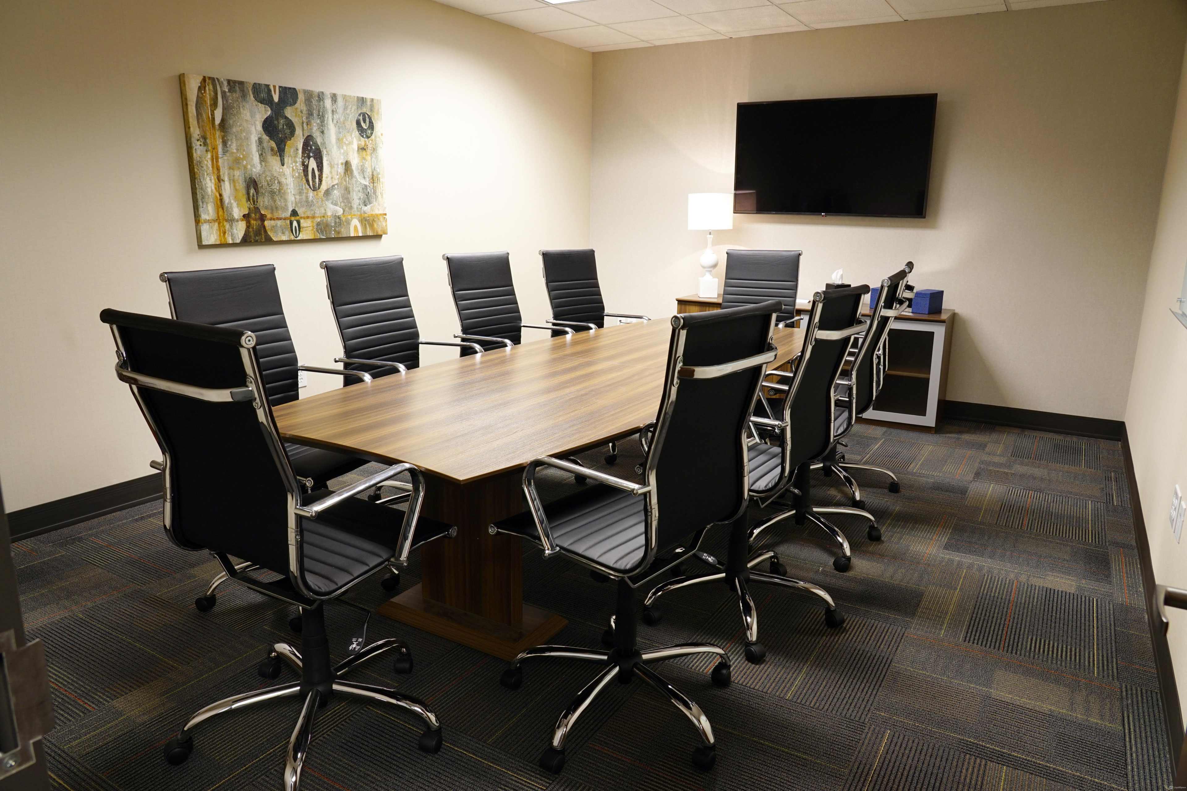 A conference room features a large wooden table surrounded by black office chairs, with a television mounted on the wall and artwork displayed.