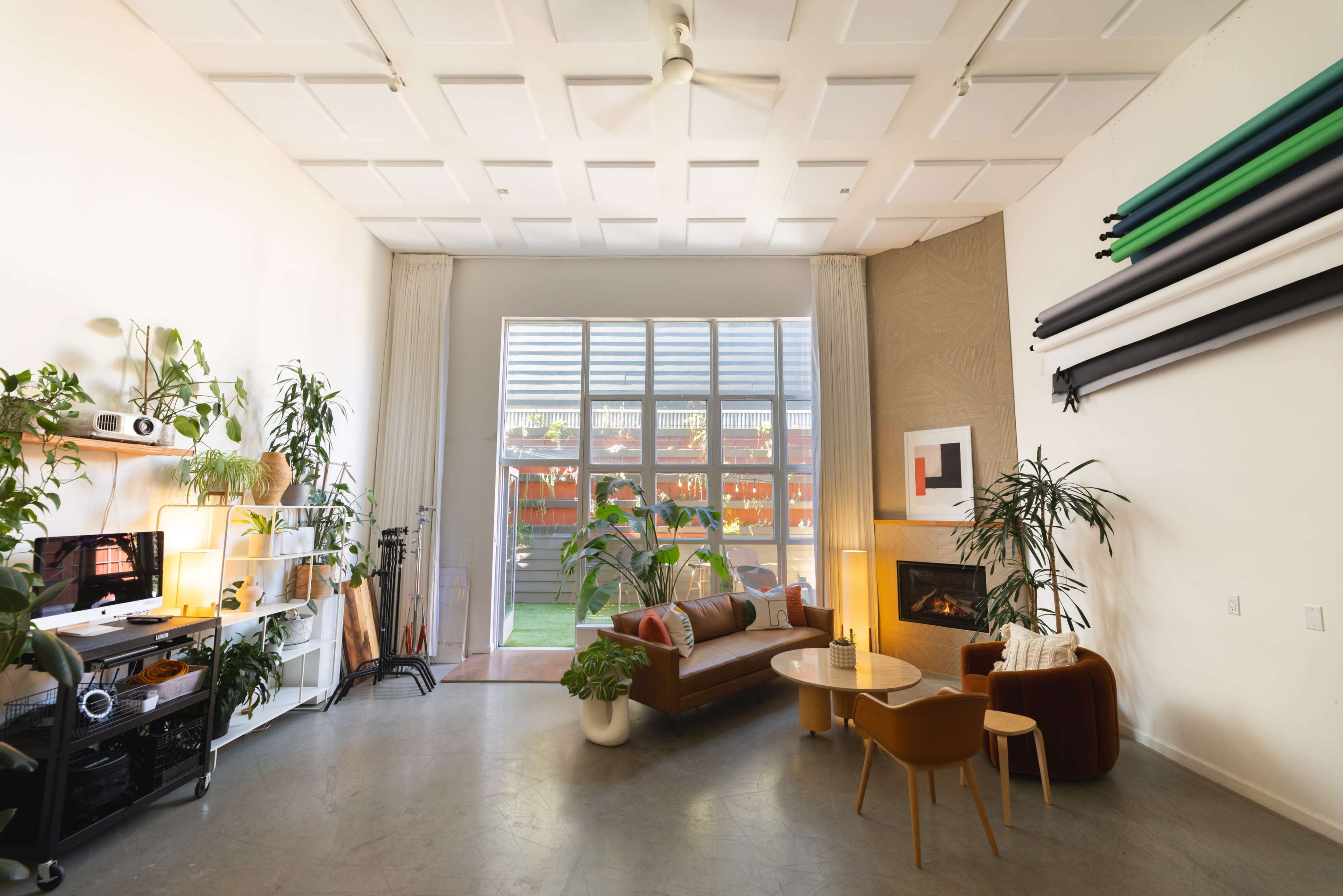 The image shows a spacious living room with large windows, a comfortable sofa, a coffee table, and various plants.