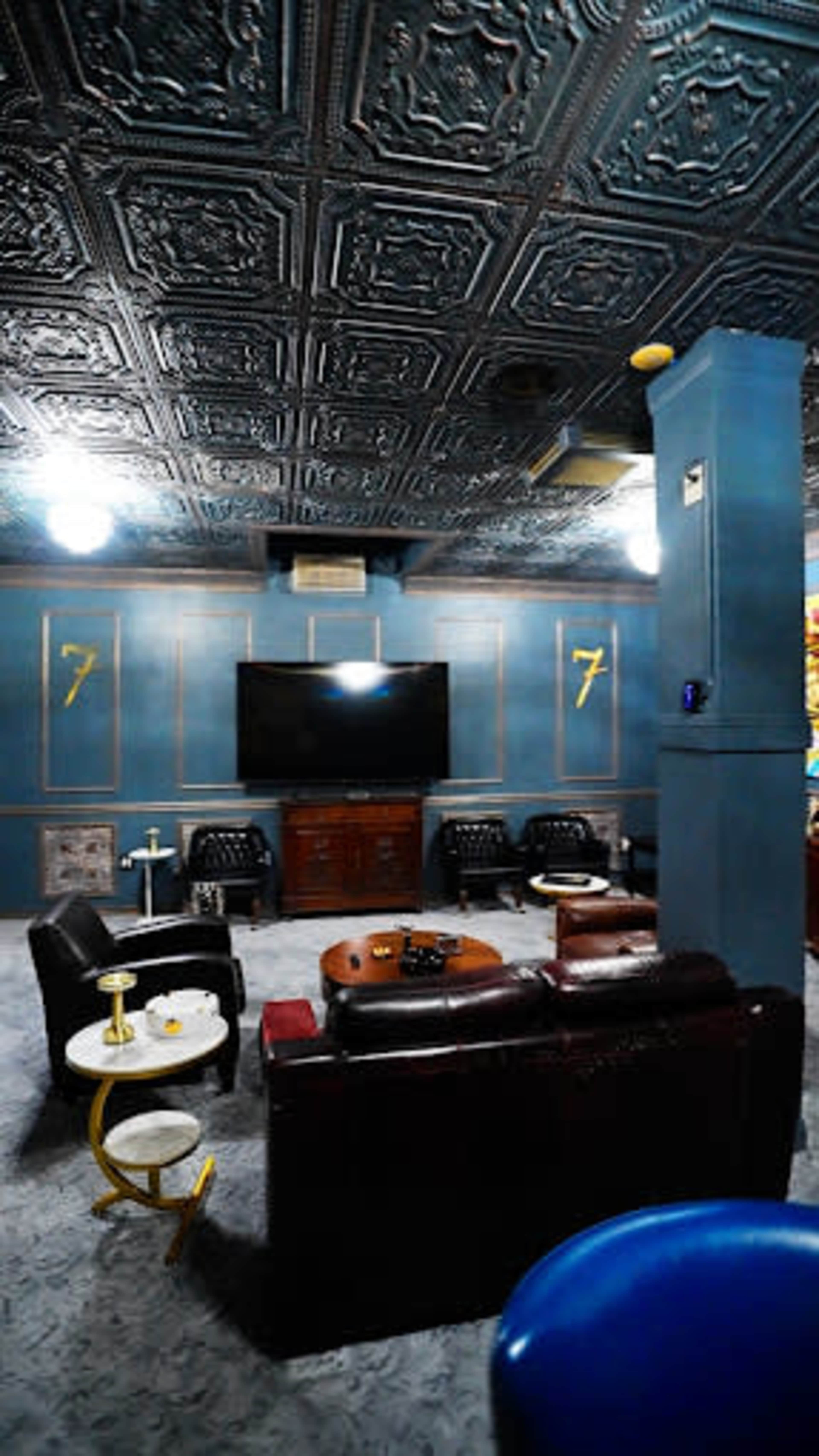 The image shows a lounge area with dark walls, a decorative ceiling, a large television, and several seating options arranged around a coffee table.