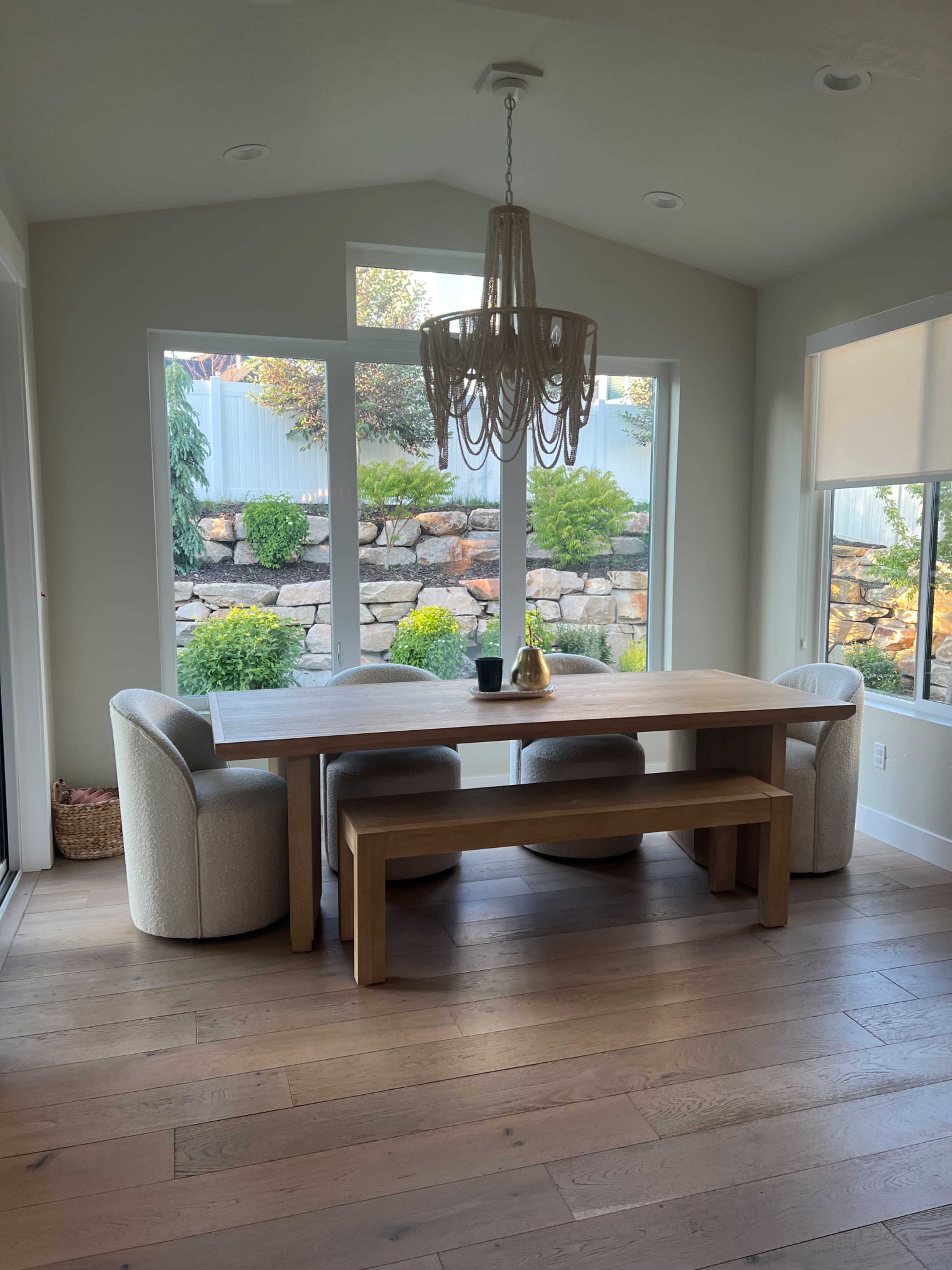 A dining area features a wooden table flanked by two curved chairs and a bench, with large windows overlooking a landscaped garden.