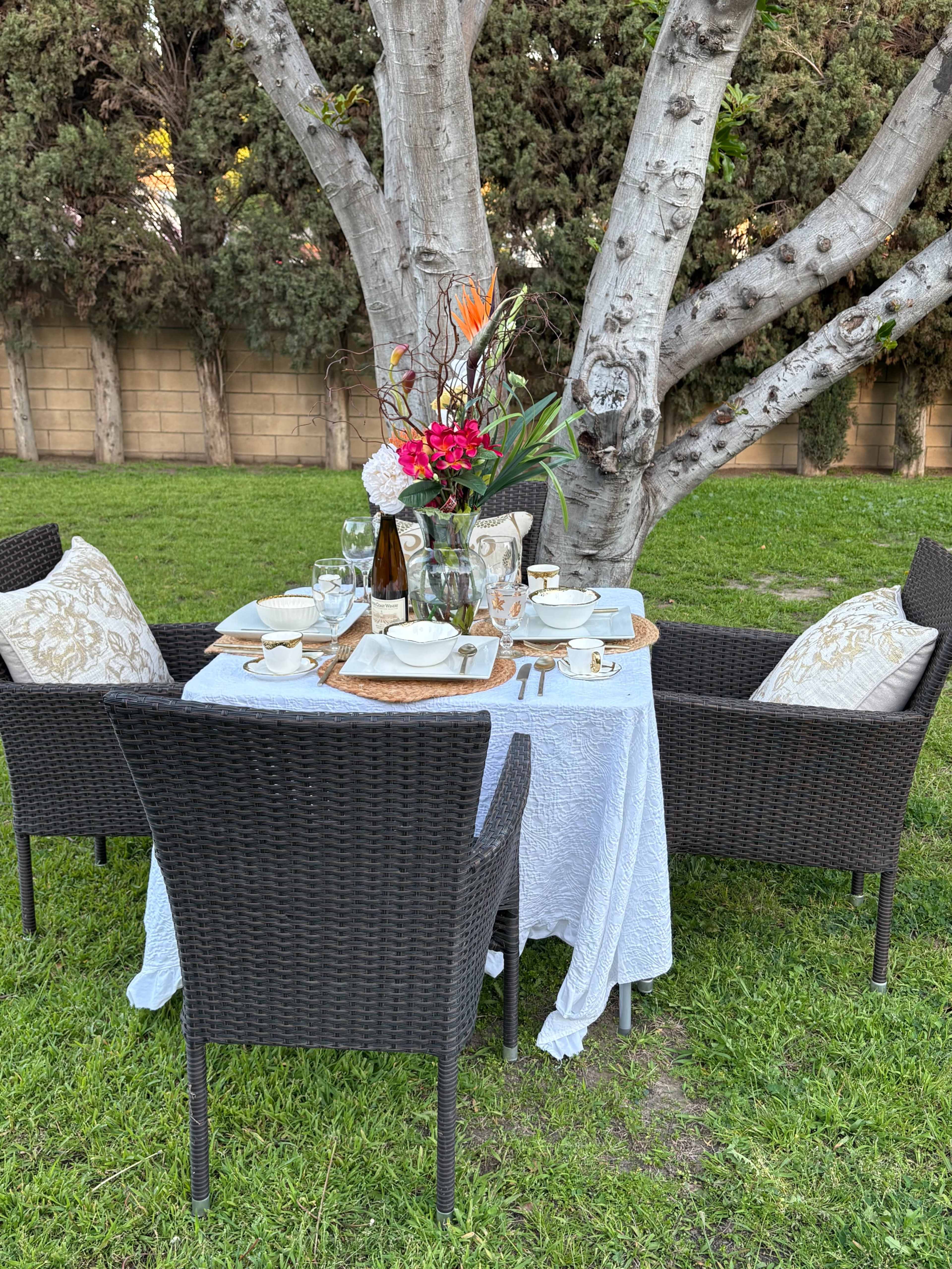 A dining table is set with floral decorations and dinnerware under a tree in a grassy outdoor area.
